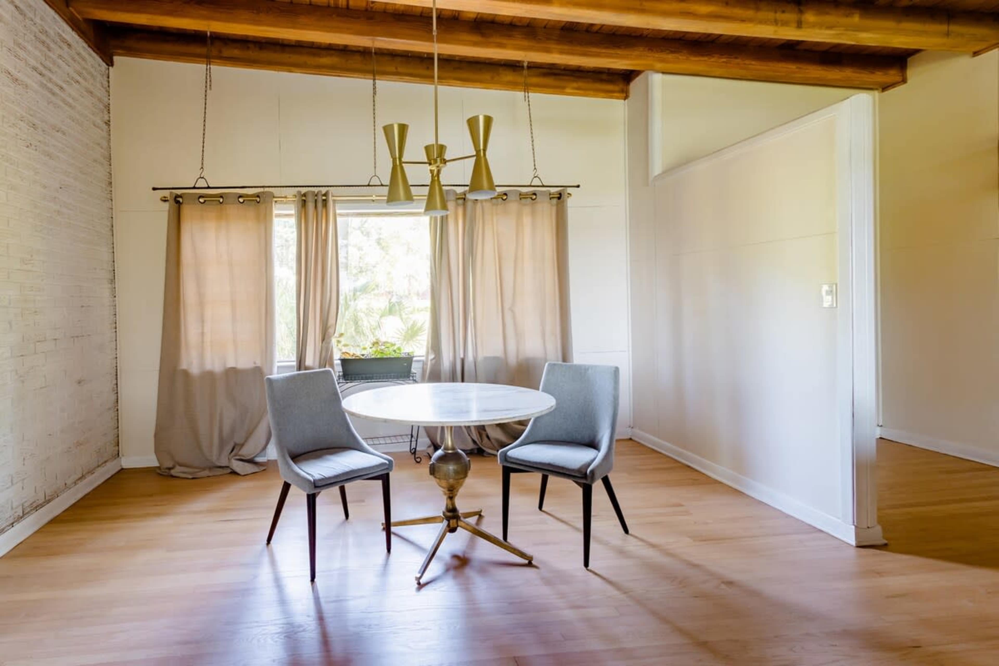 A circular table with two chairs is positioned beneath a window with light curtains in a room featuring wooden beams and hardwood floors.