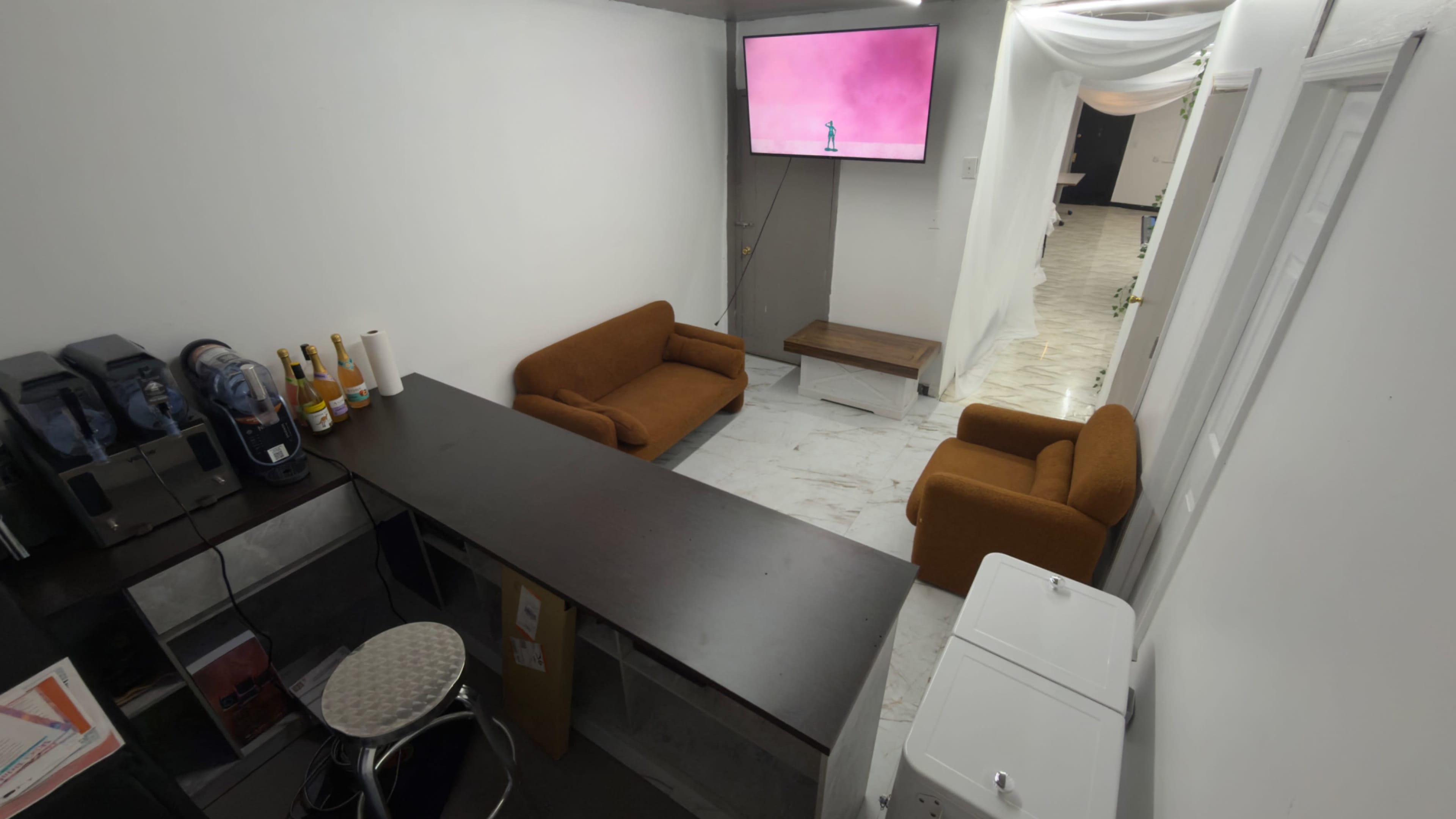 The image shows a small, modern lounge area with two orange sofas, a black countertop, and a wall-mounted television displaying a pink screen.