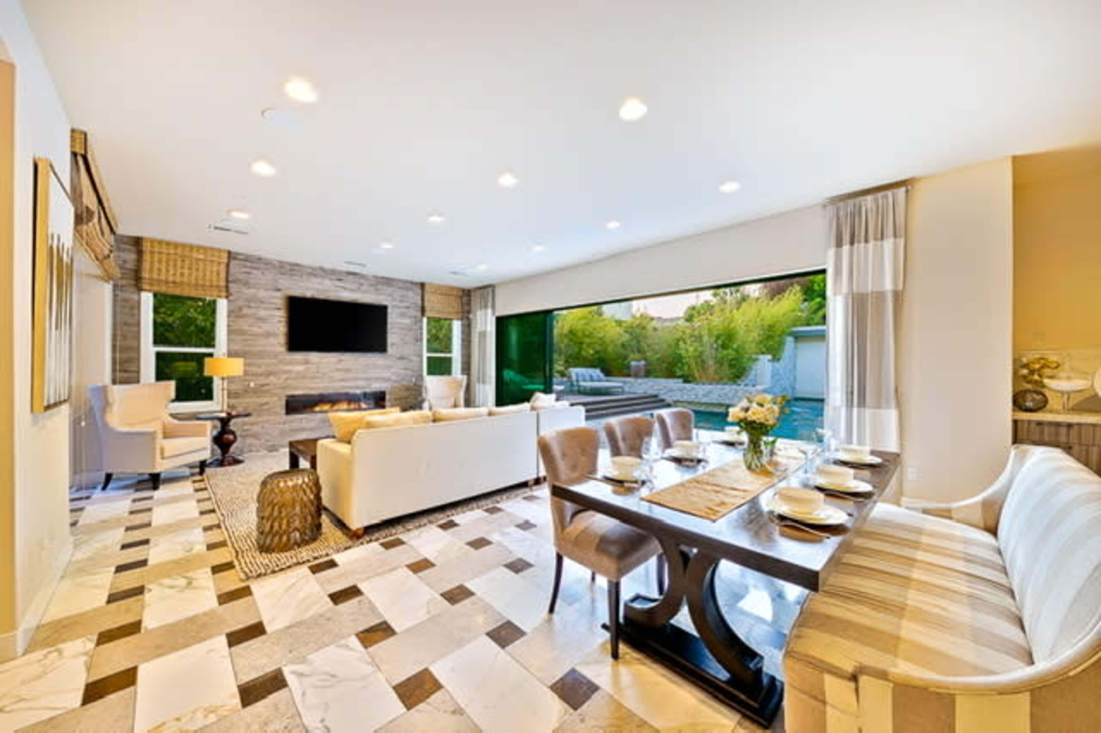 A modern living and dining area features a neutral color scheme, large windows overlooking a garden, and a dining table set for a meal.