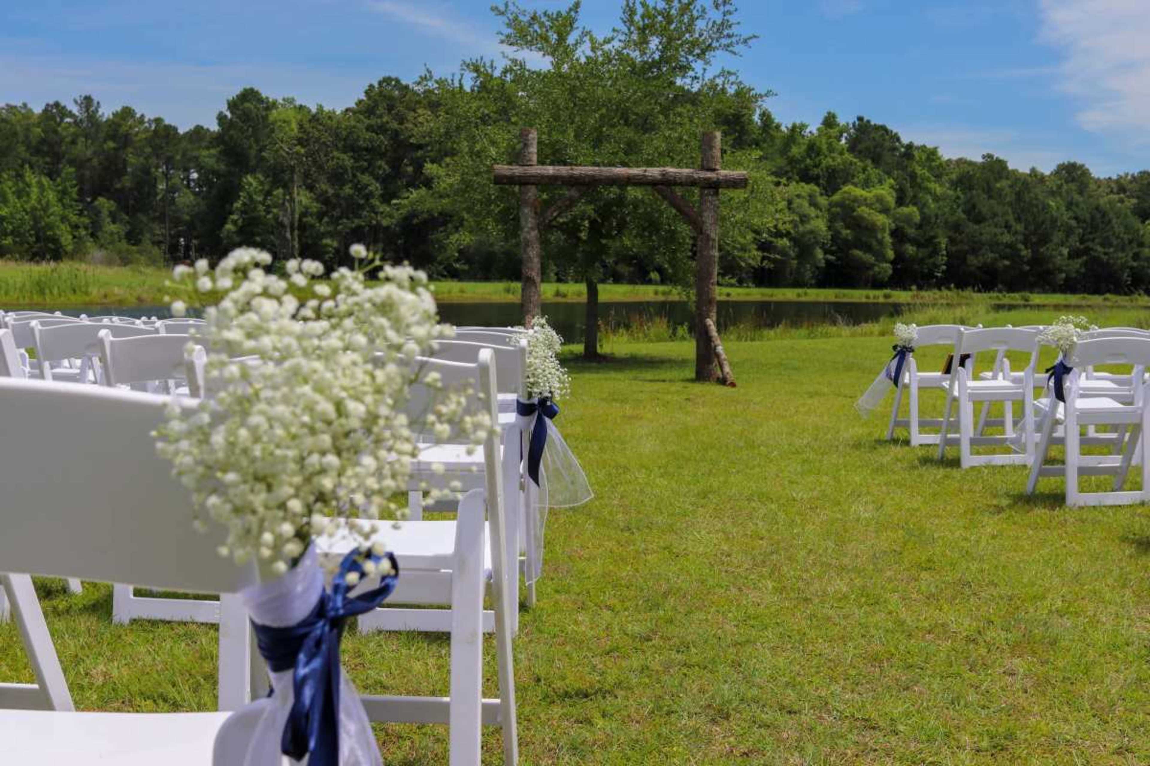 An outdoor wedding setup features white chairs adorned with baby’s breath bouquets and blue ribbons, facing a wooden arch surrounded by greenery and a pond.