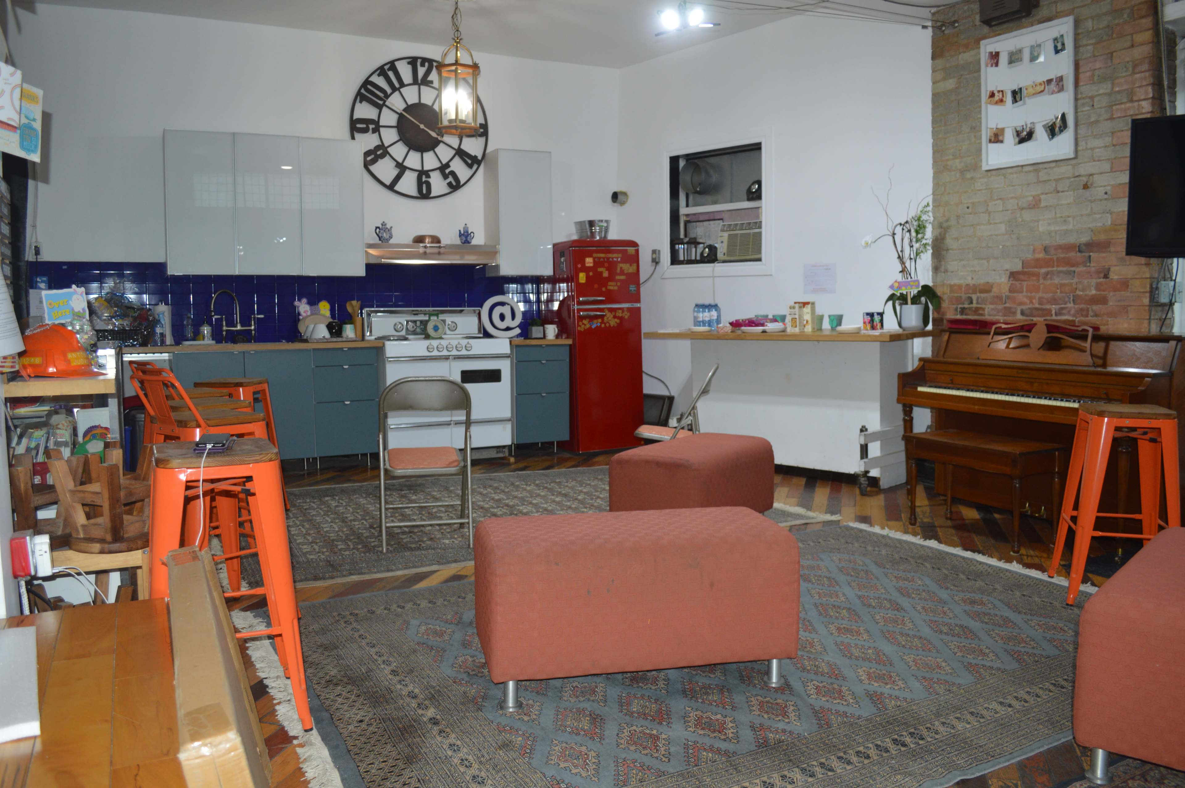 The image shows a cozy kitchen and living area featuring a blue-tiled counter with a vintage stove, bright orange stools, two orange armchairs on a patterned rug, and a piano against a brick wall.