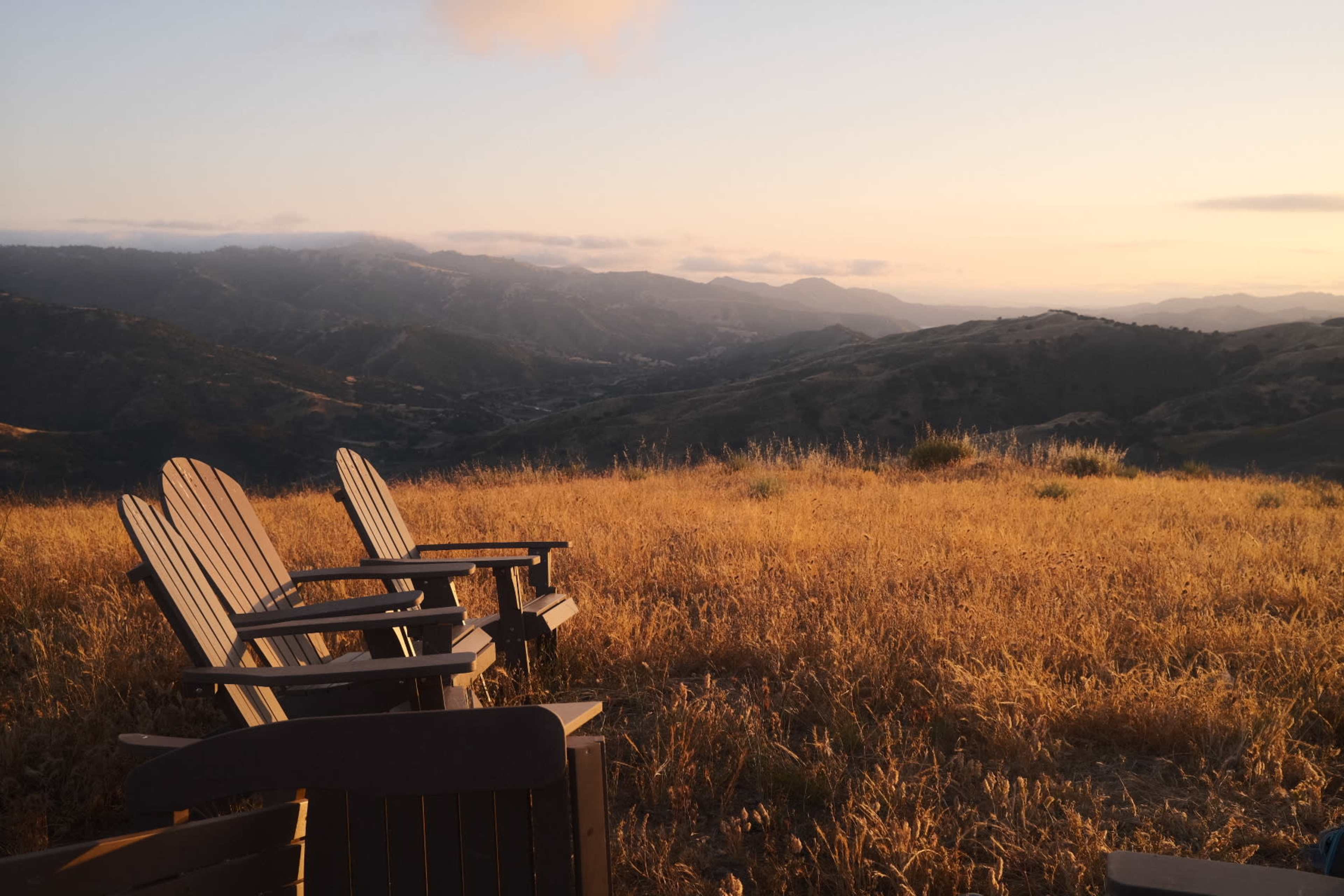 A row of wooden Adirondack chairs facing a panoramic view of rolling hills and mountains at sunset.