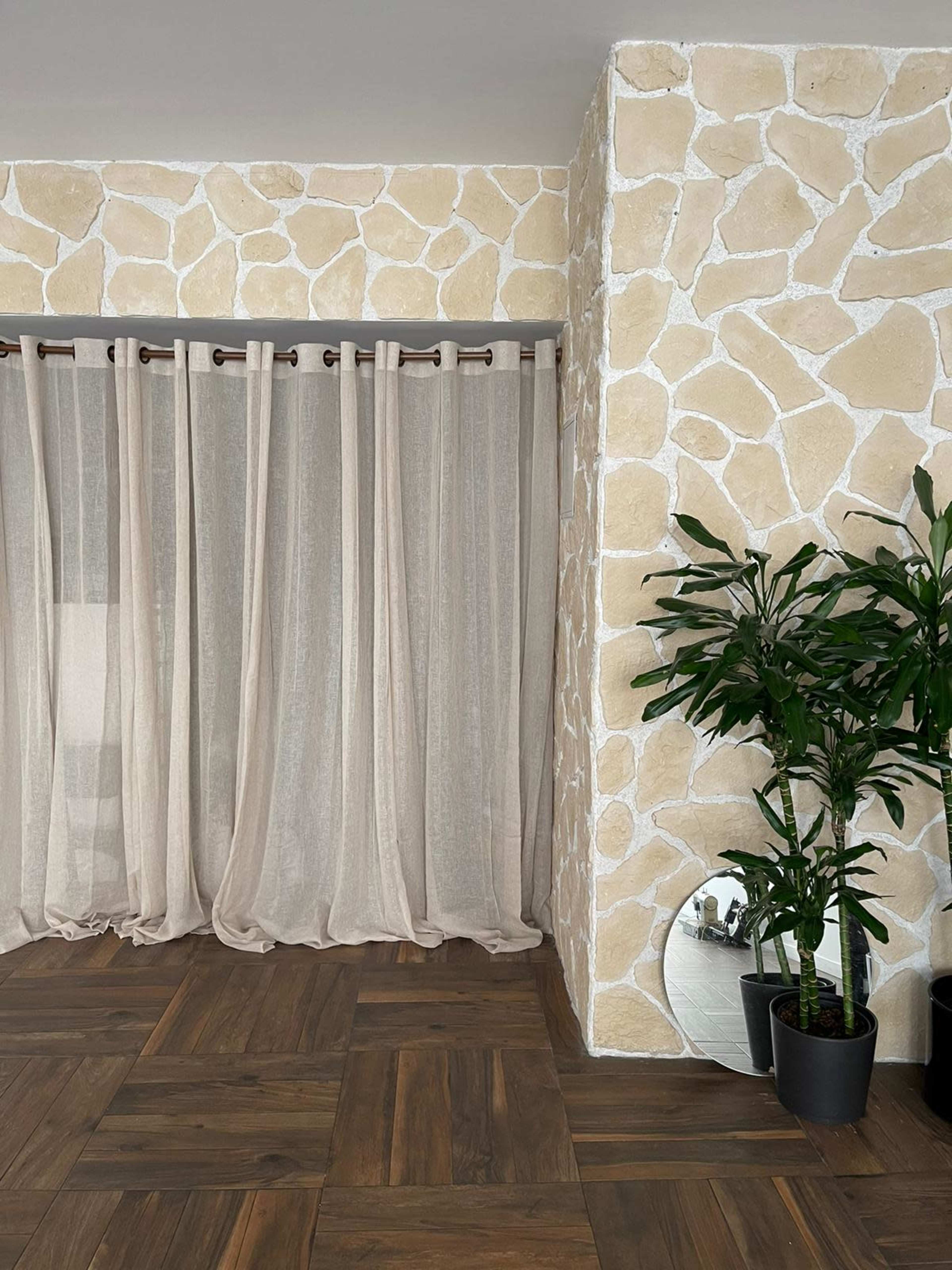 A room with a stone-textured wall, sheer curtains, a potted plant, and a round mirror against a wooden floor.