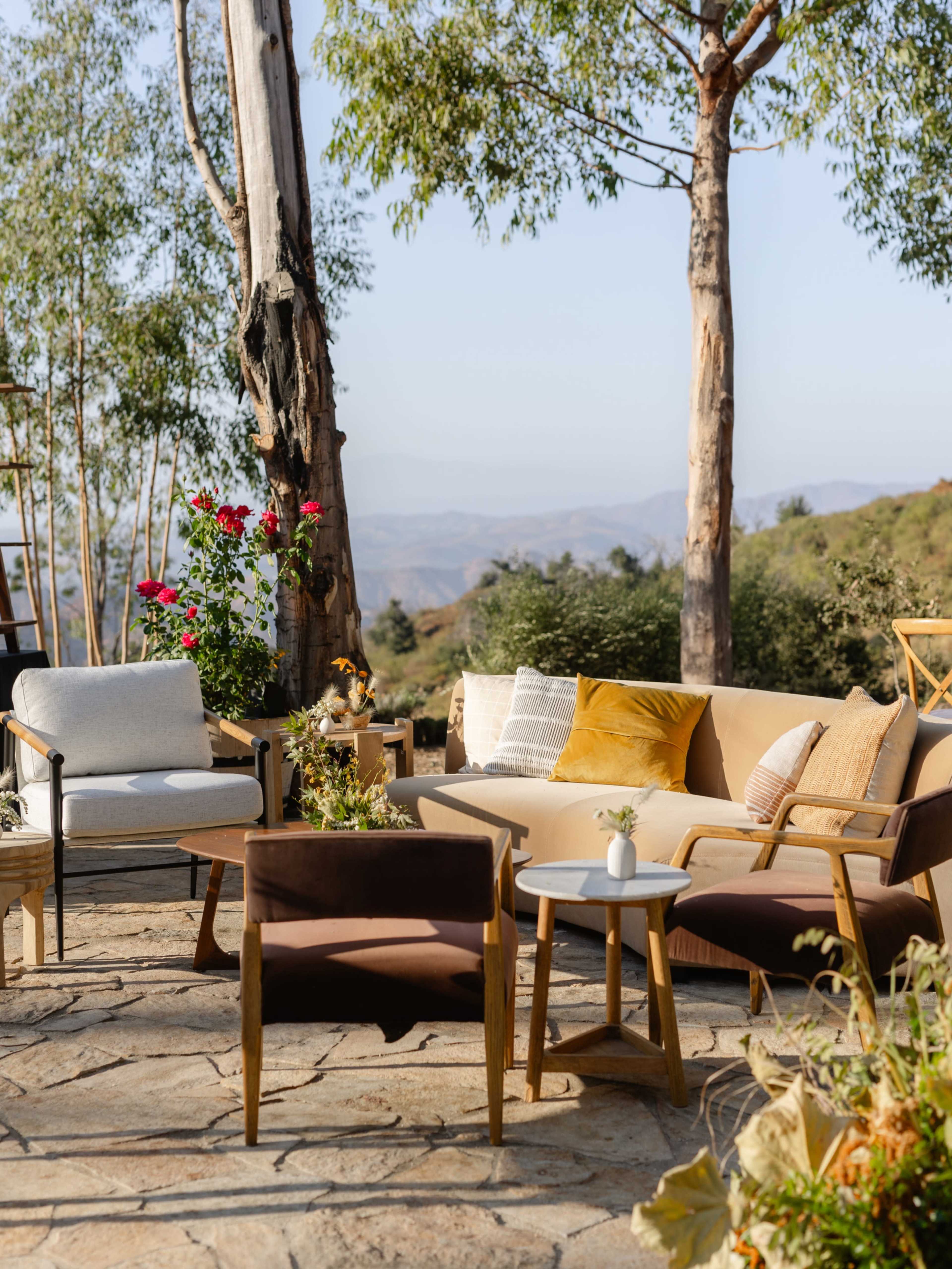 The scene features a well-arranged outdoor seating area with various chairs and sofas set on a stone patio, surrounded by trees and distant hills.