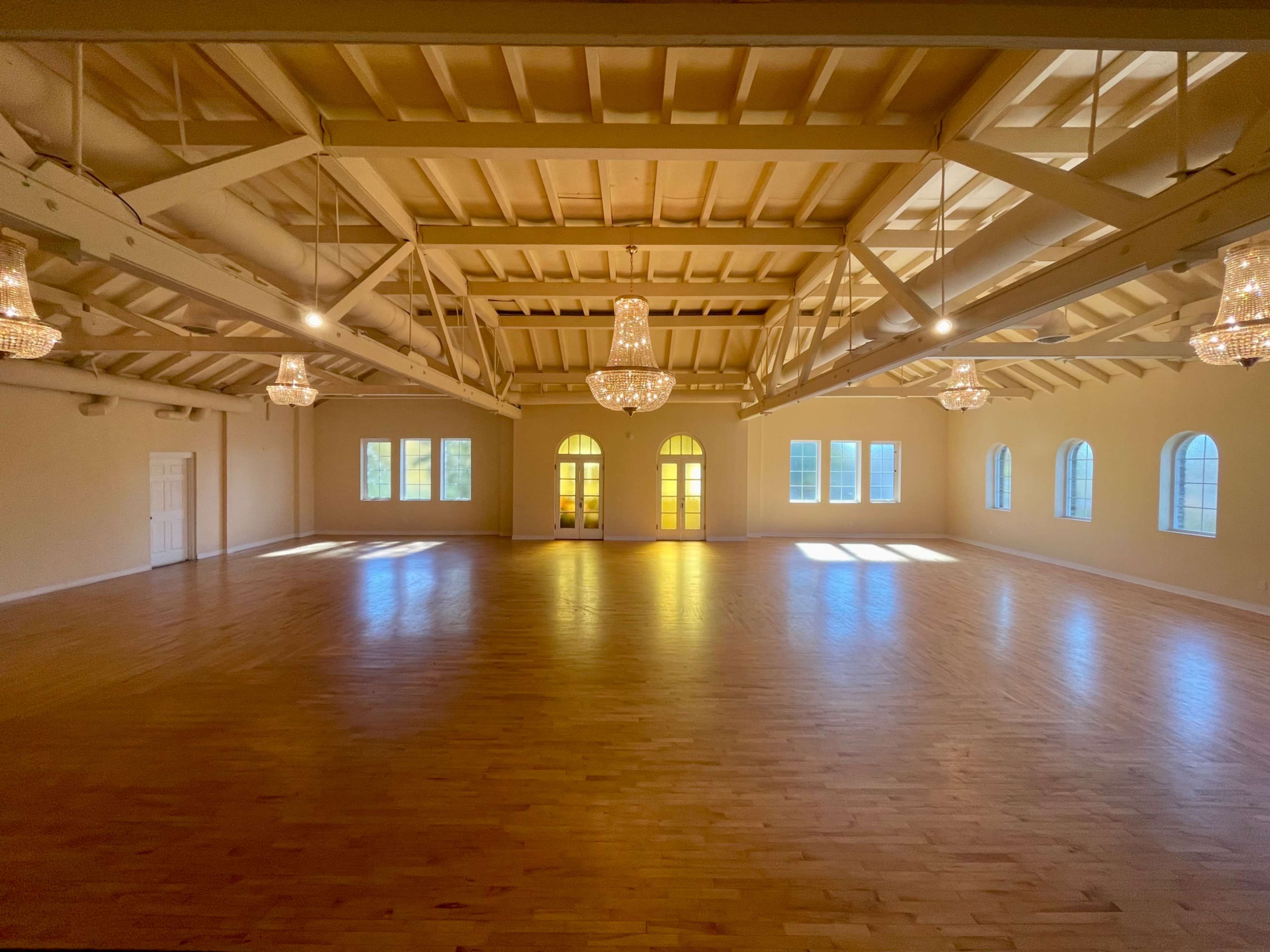 The image shows a spacious, well-lit room with wooden floors, high ceilings, and several chandeliers, featuring arched windows and double doors at one end.