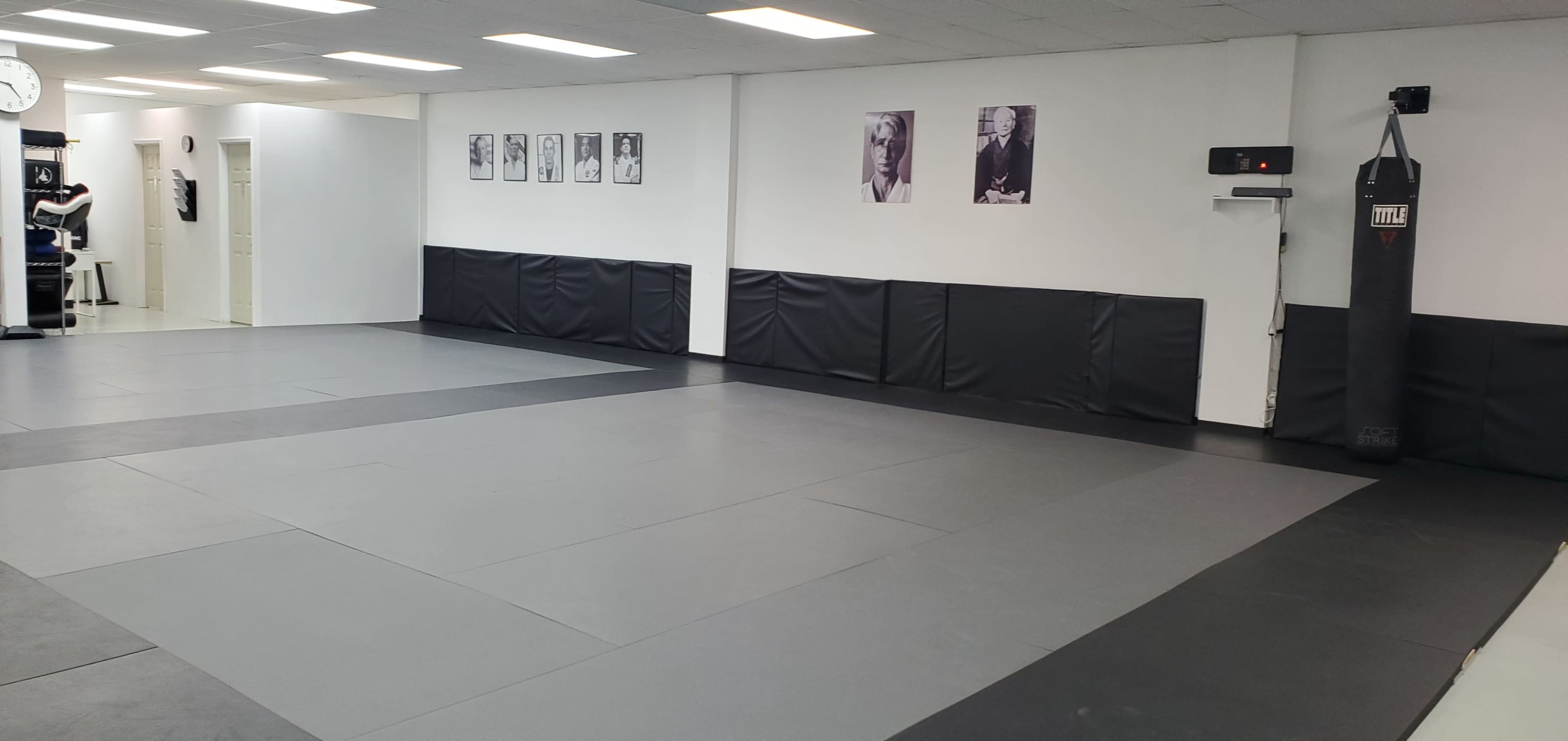A spacious martial arts gym with black and gray mats, black wall padding, and framed photographs on the walls.