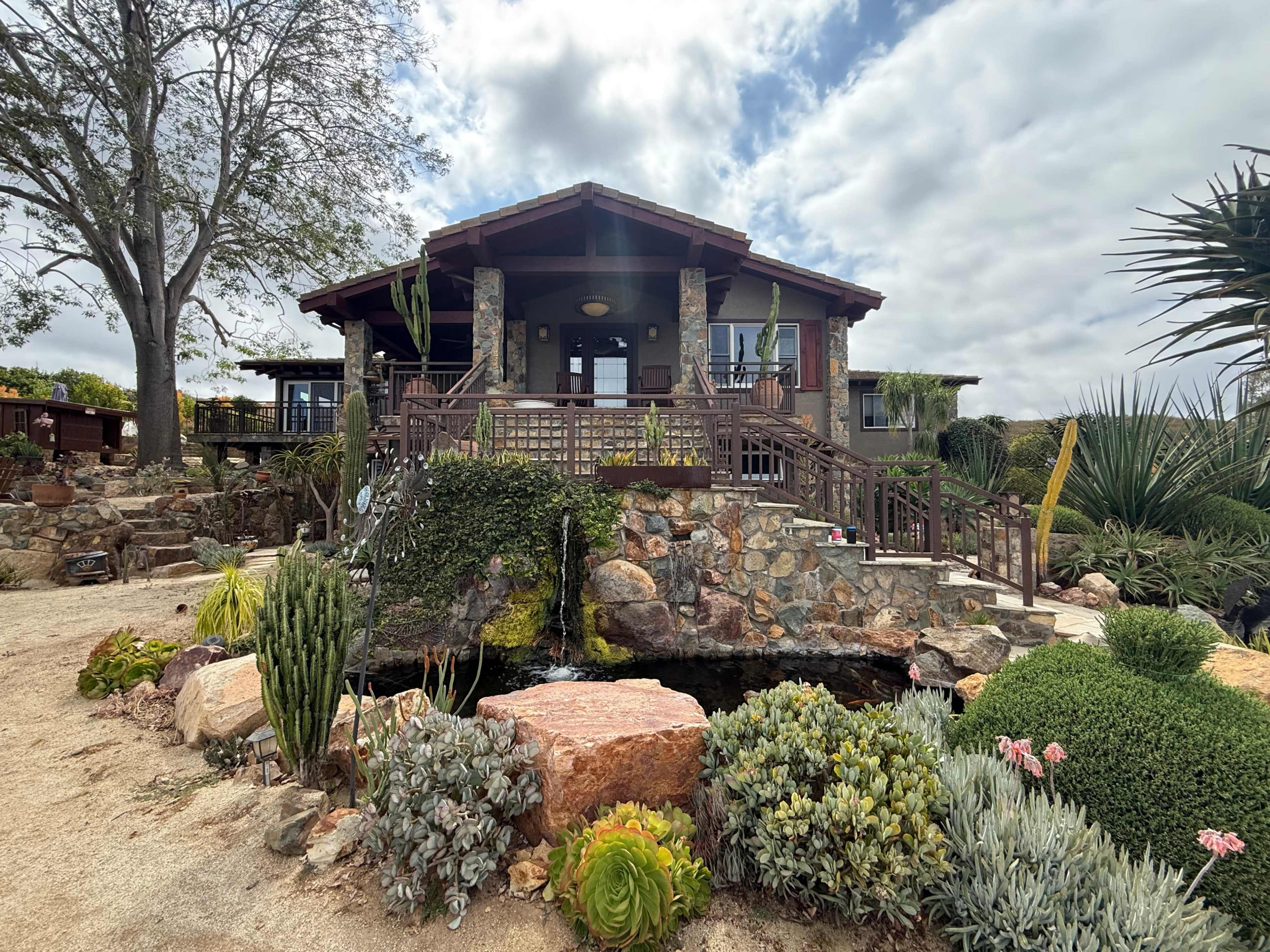The image shows a rustic-style house surrounded by lush greenery and various types of plants, with a small pond and landscaped rocky features in the foreground.