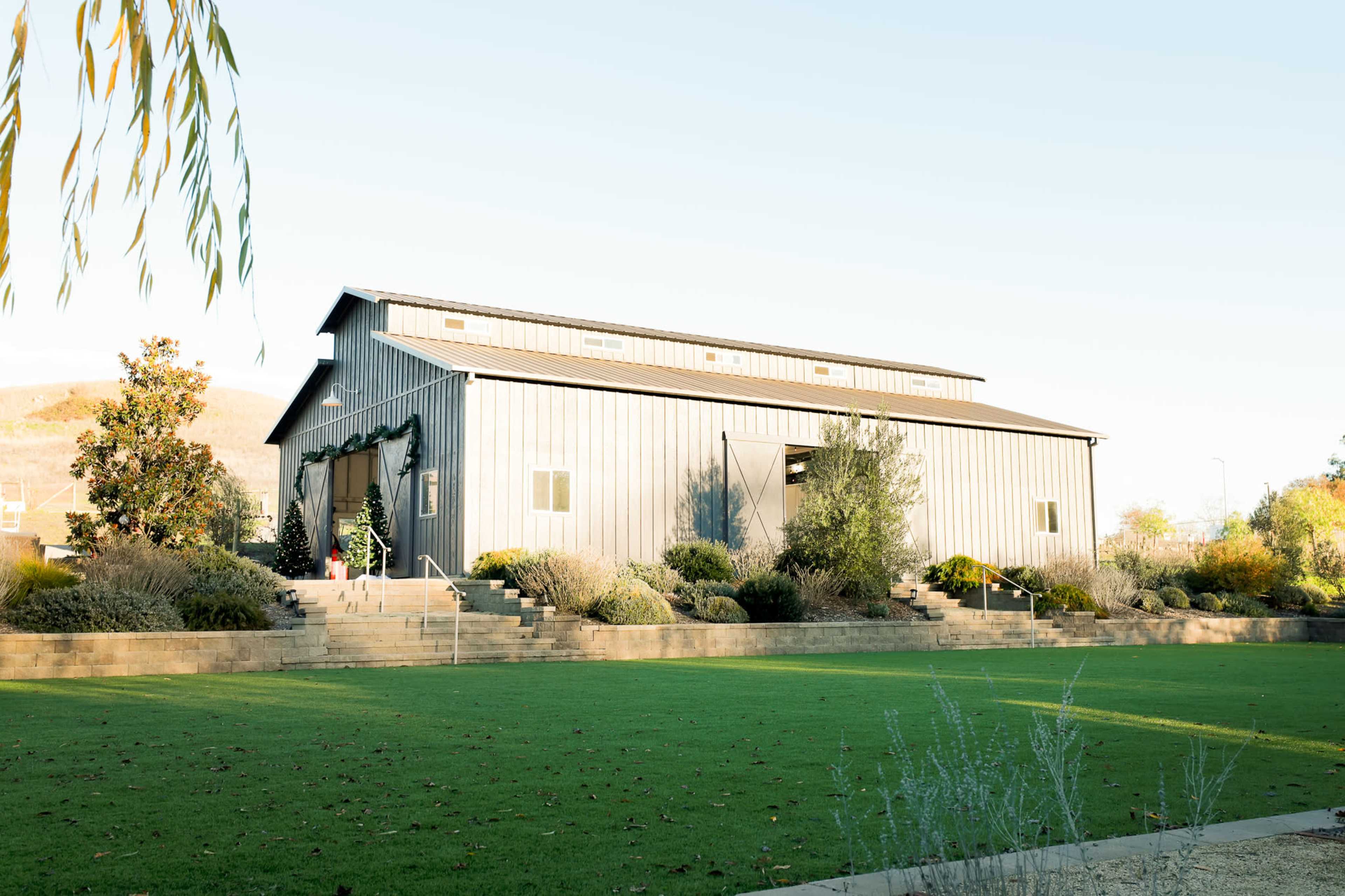 A large, gray metal barn is set on landscaped grounds with steps leading up to it.