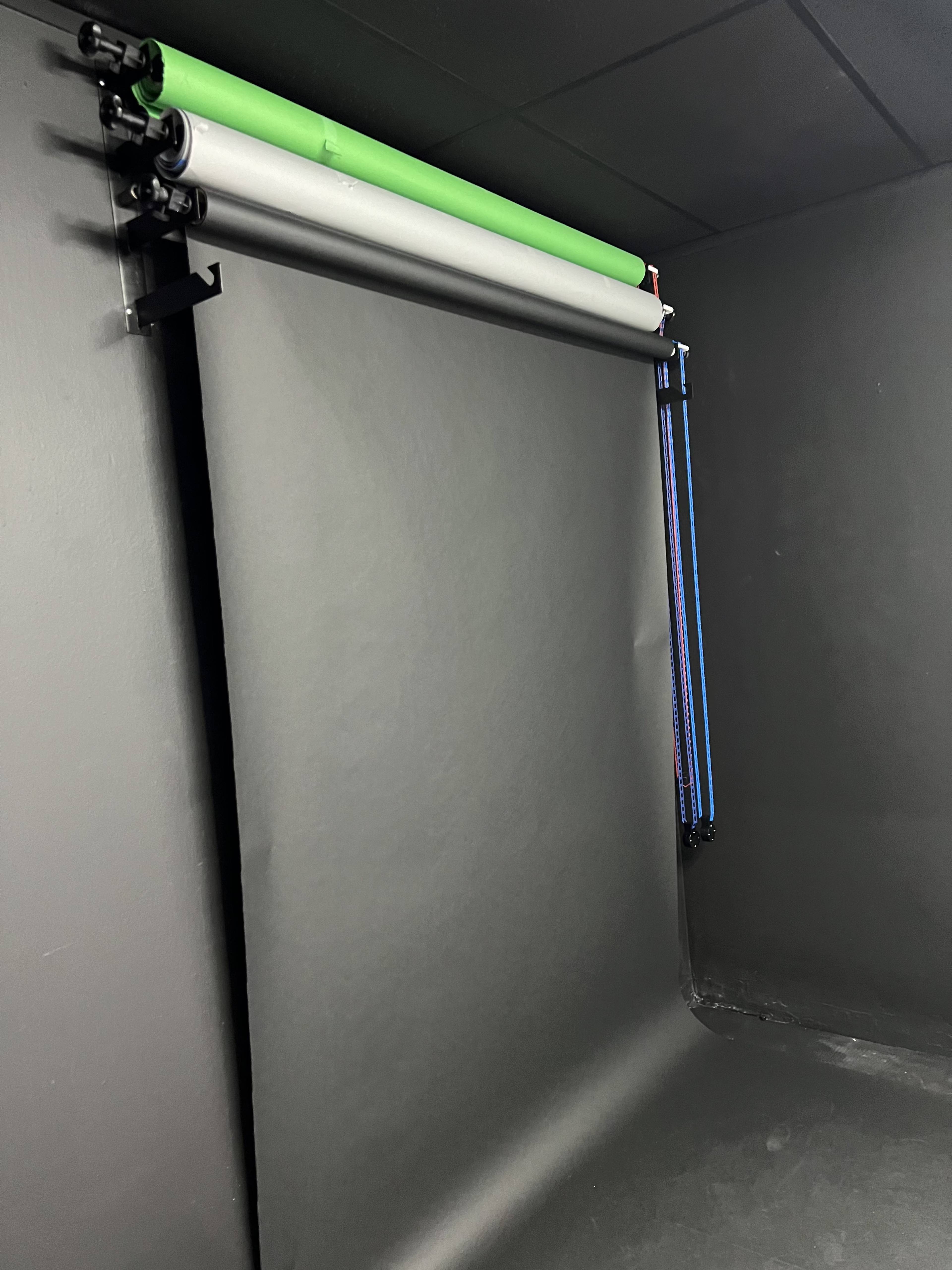 The image shows a setup for photography or videography with a backdrop system featuring rolled background materials in green, gray, and blue, mounted on a wall.