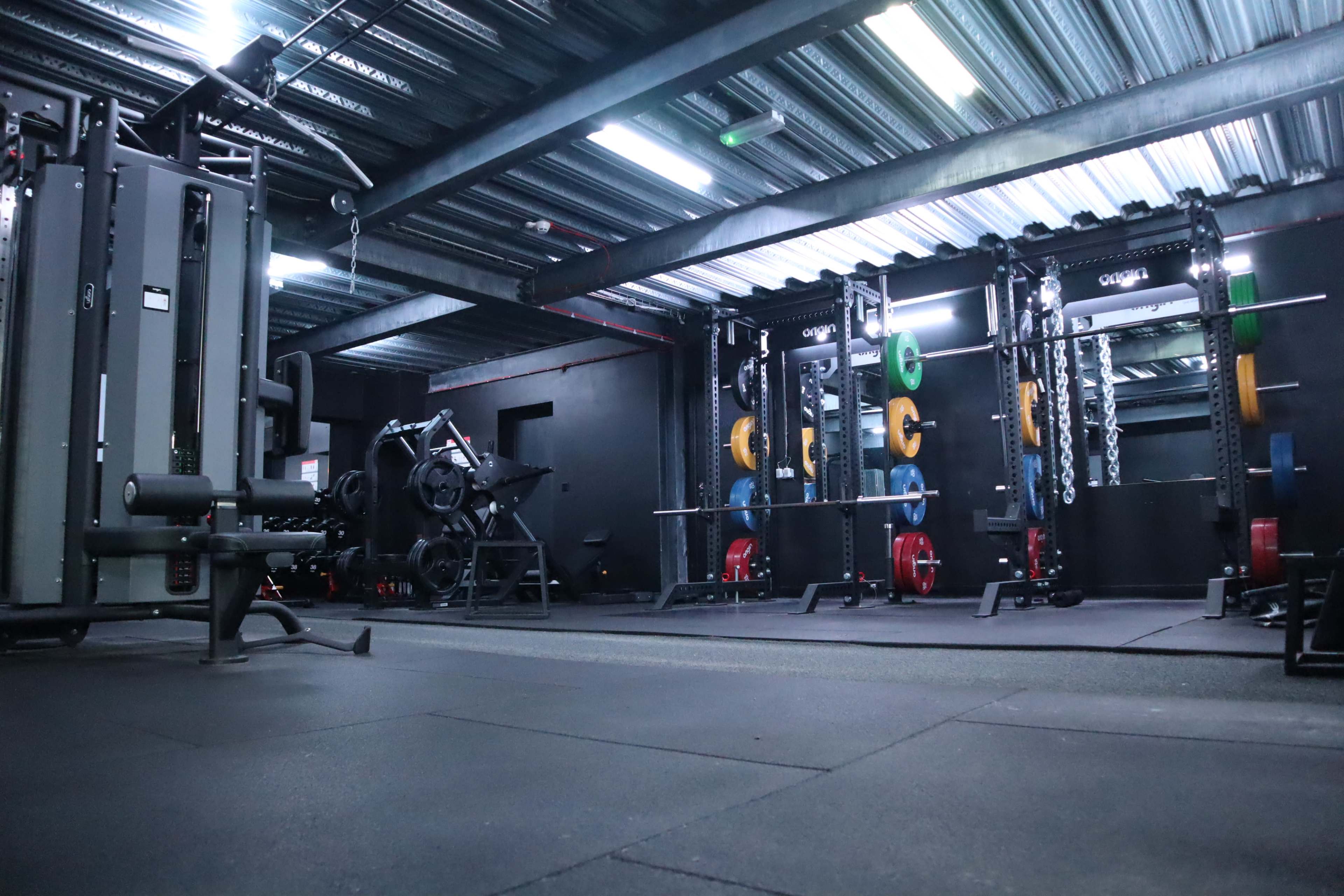 The image shows a dimly lit gym interior with weightlifting equipment and benches arranged along the walls.