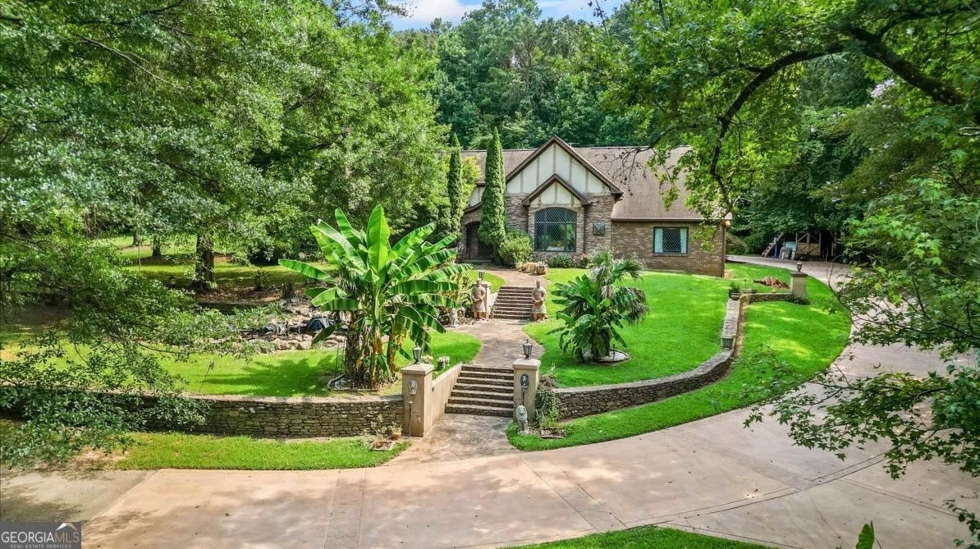 A large, two-story brick house with a gable roof is situated on a spacious, landscaped lot, featuring a circular driveway and lush greenery surrounding it.
