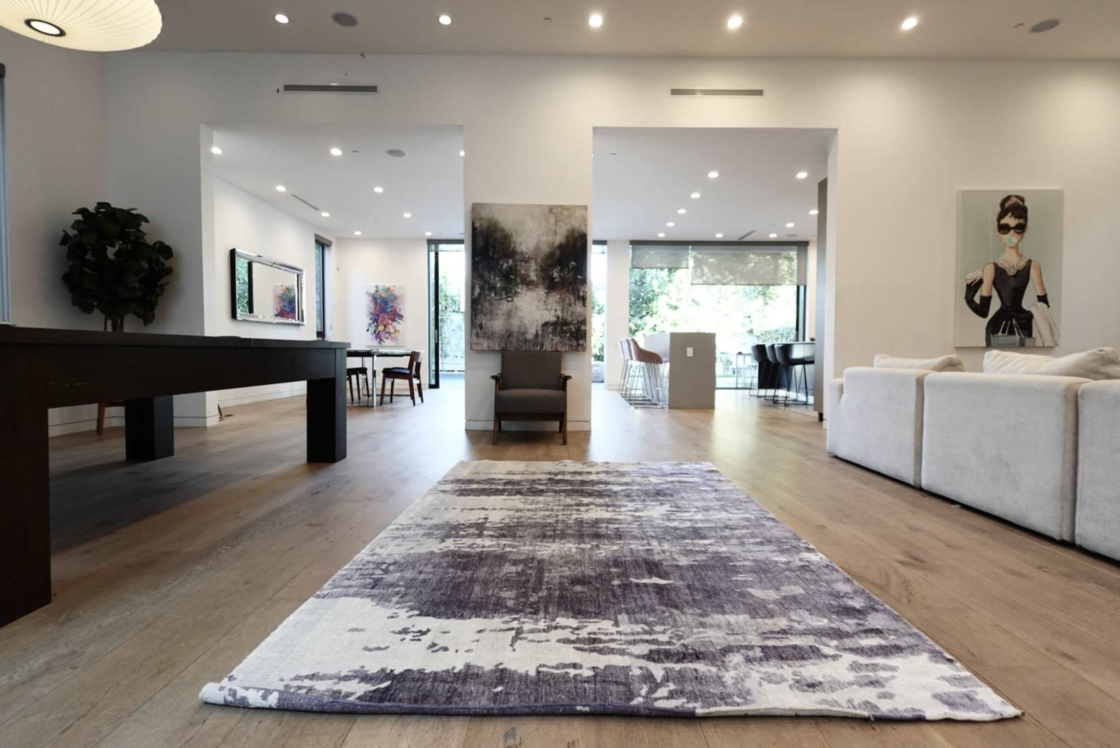 A spacious, modern living area features a large rug, a seating arrangement, a dining space, and large windows allowing natural light to enter.