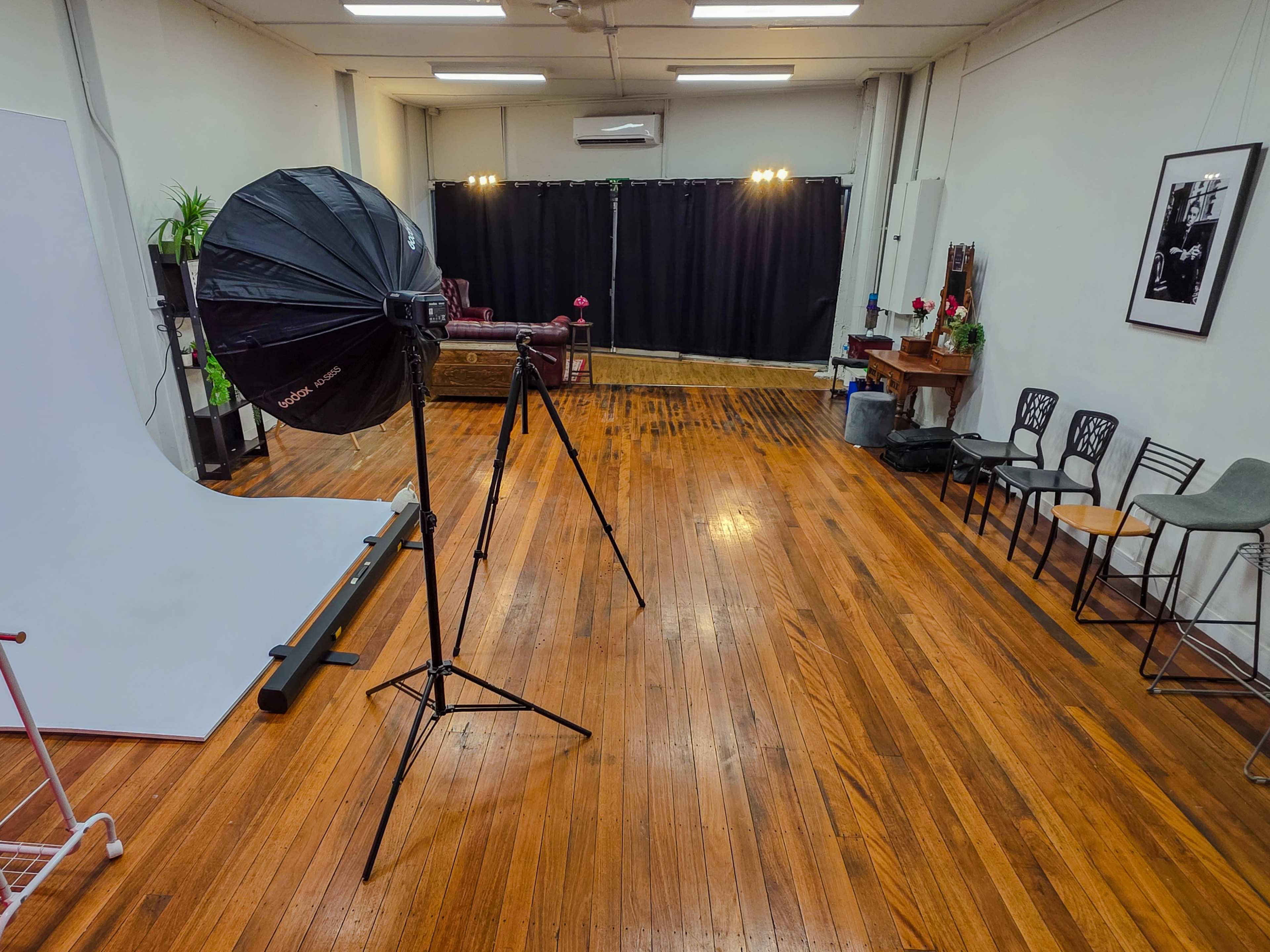 The image shows a spacious photography studio with wooden floors, a backdrop setup, a lighting umbrella on a tripod, and chairs arranged along one side.