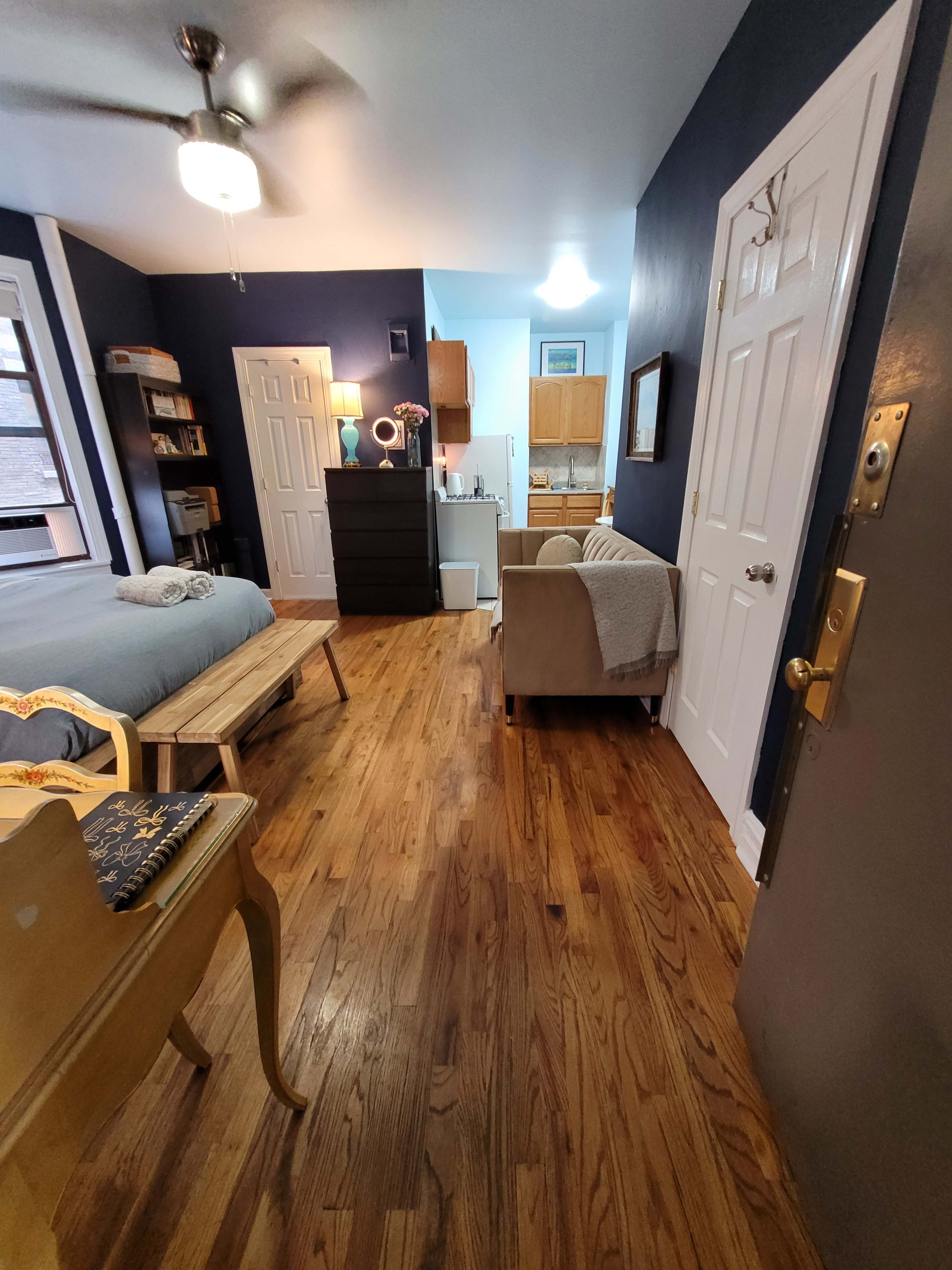 A small studio apartment features a bed on the left, a seating area with a couch on the right, and a kitchenette visible in the background, all with wooden floors and blue walls.