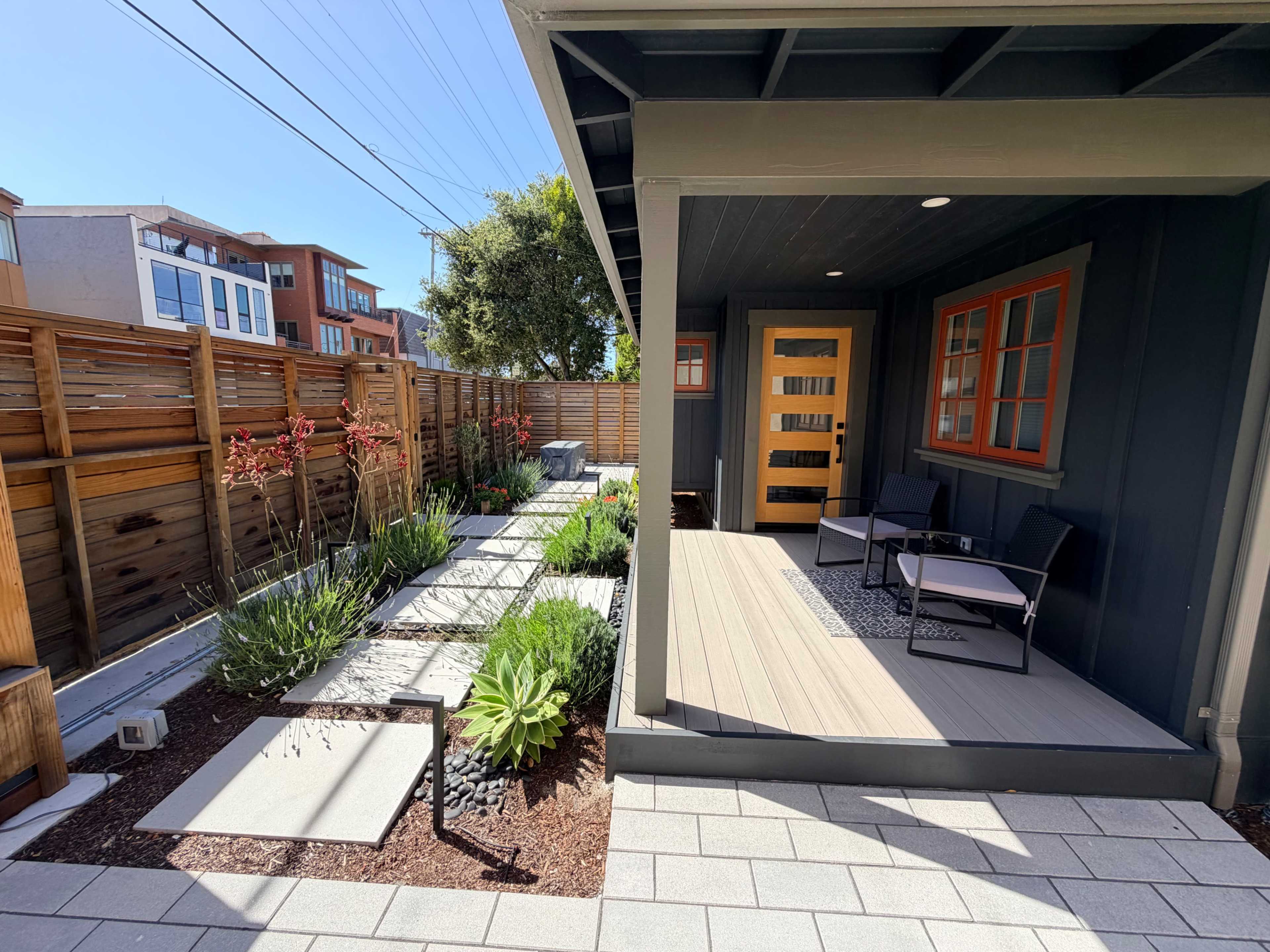 The image shows a modern outdoor patio area adjacent to a landscaped garden featuring stone walkways and various plants.