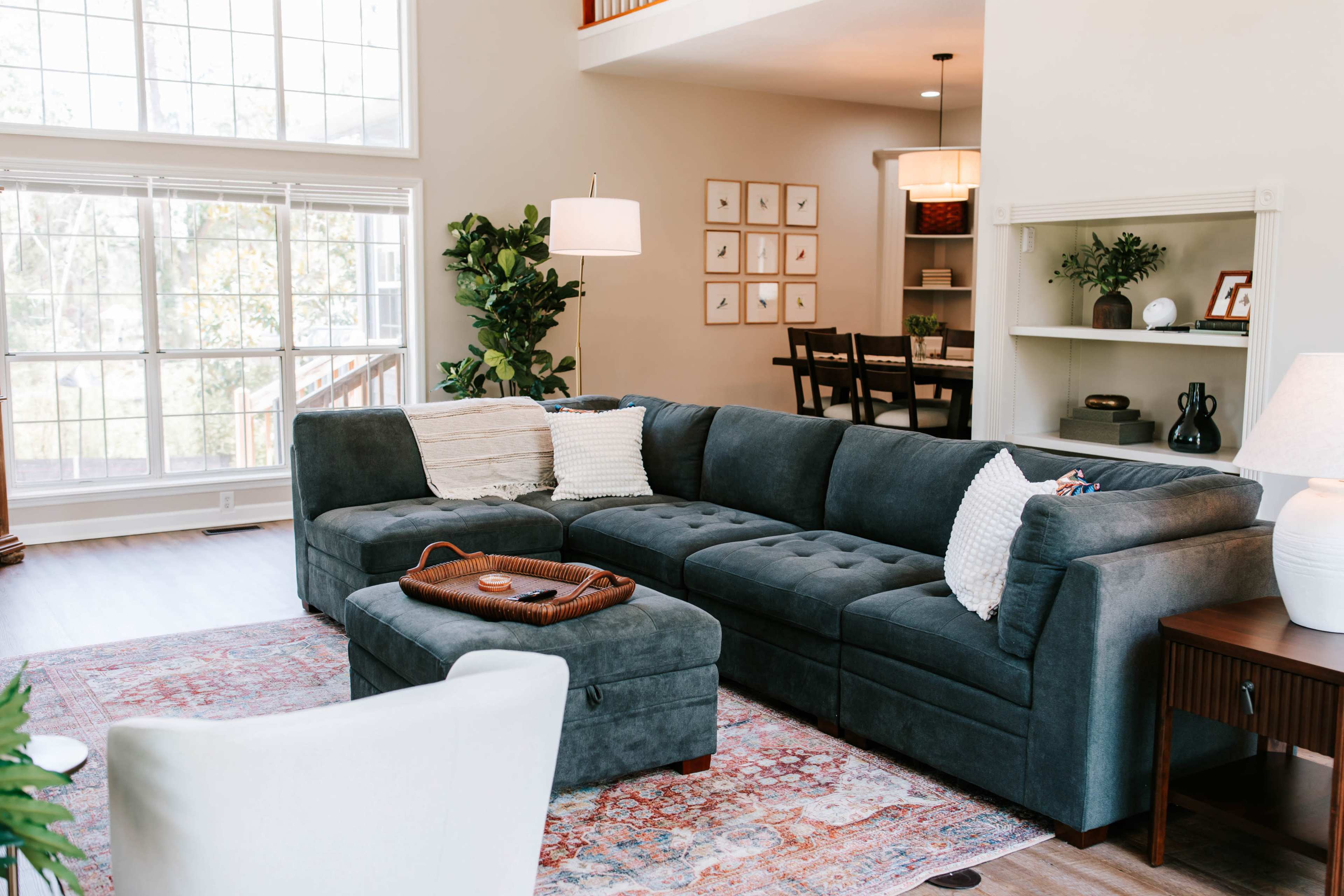 The image shows a modern living room featuring a large grey sectional sofa, a small ottoman, and large windows that allow natural light to illuminate the space.
