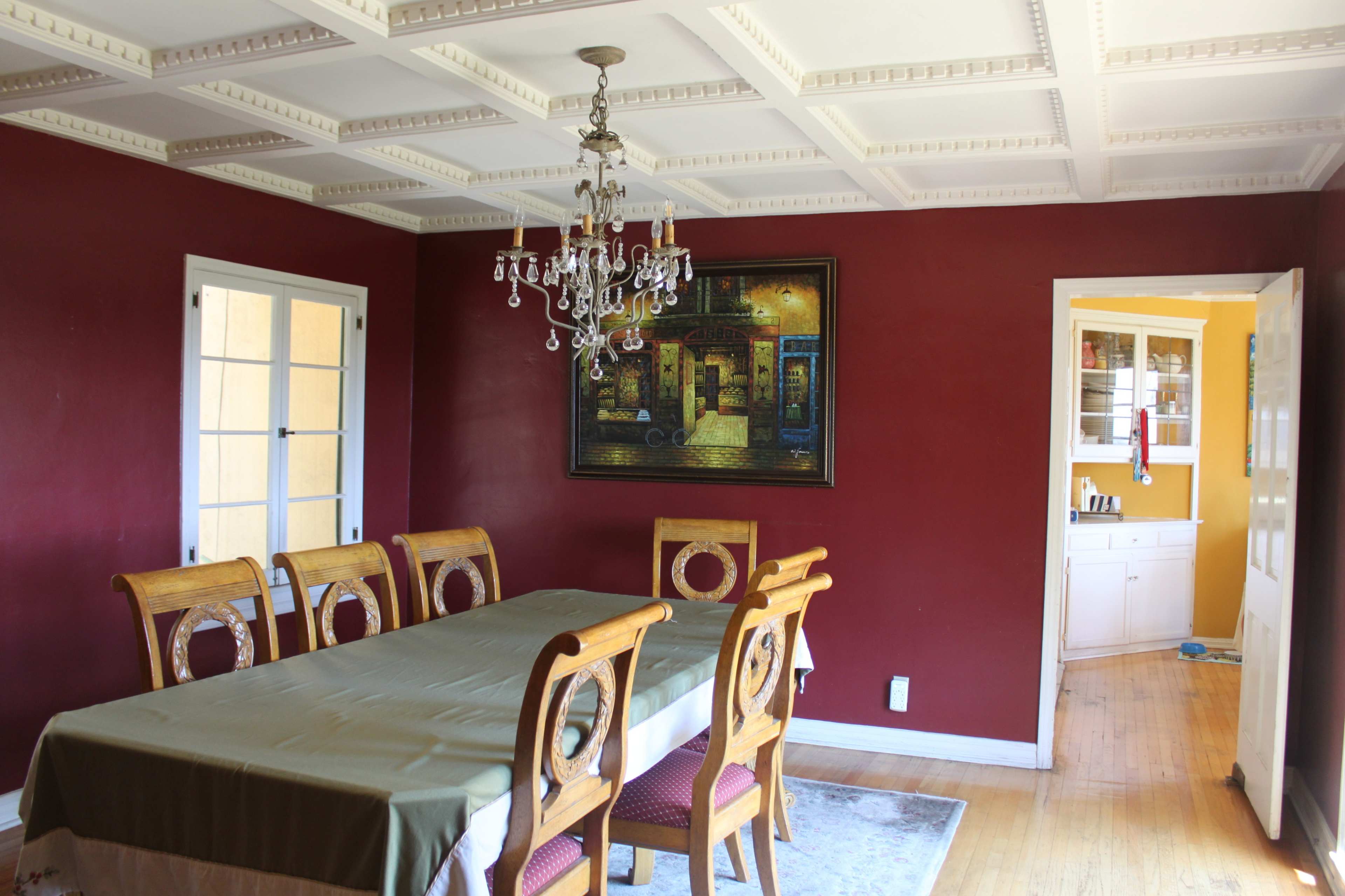 A dining room features a long table surrounded by wooden chairs, with a chandelier hanging overhead and a painting on the red wall.