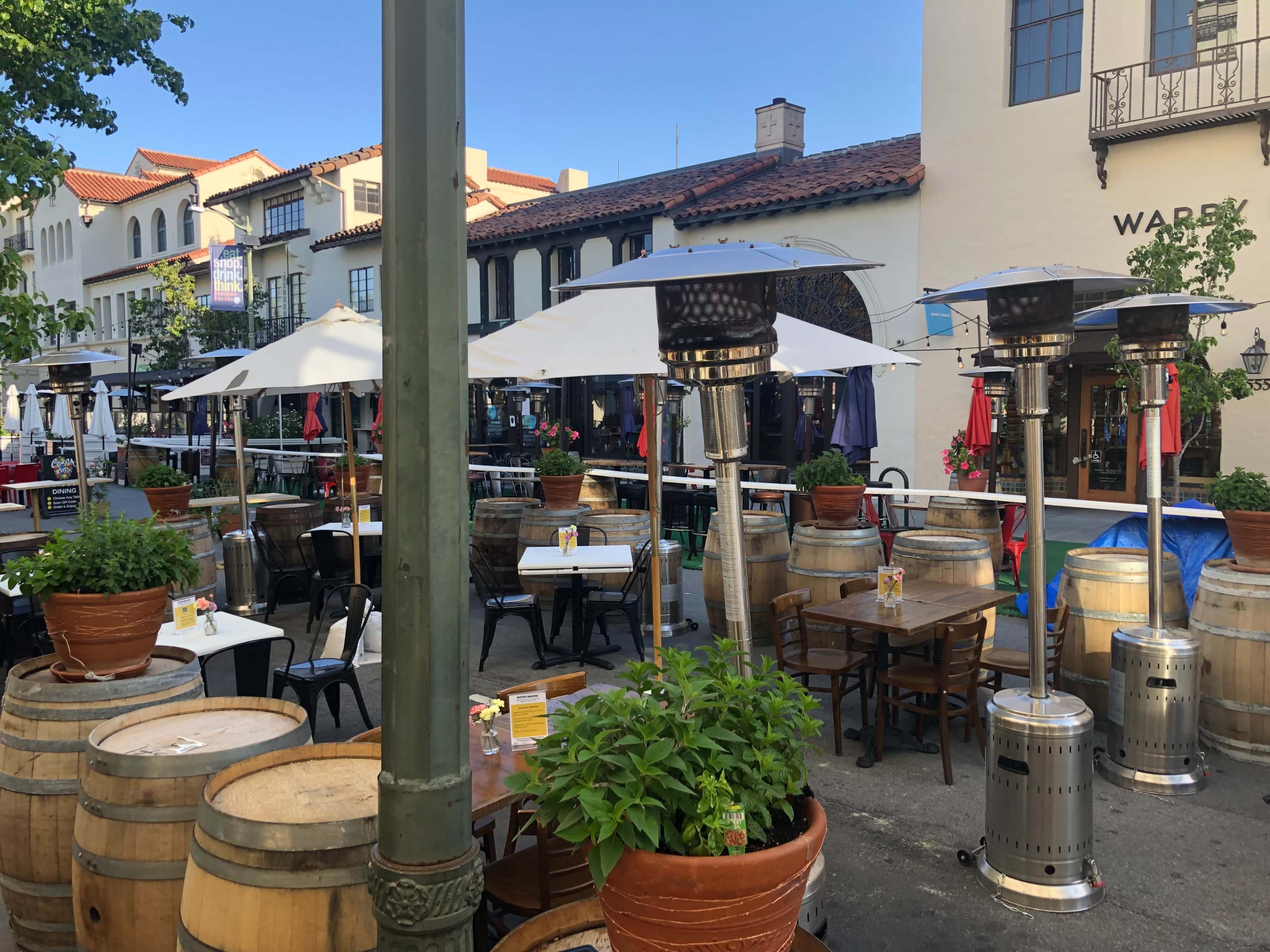 A patio area with tables and chairs surrounded by potted plants and umbrellas, featuring outdoor heaters and wine barrels as decoration.