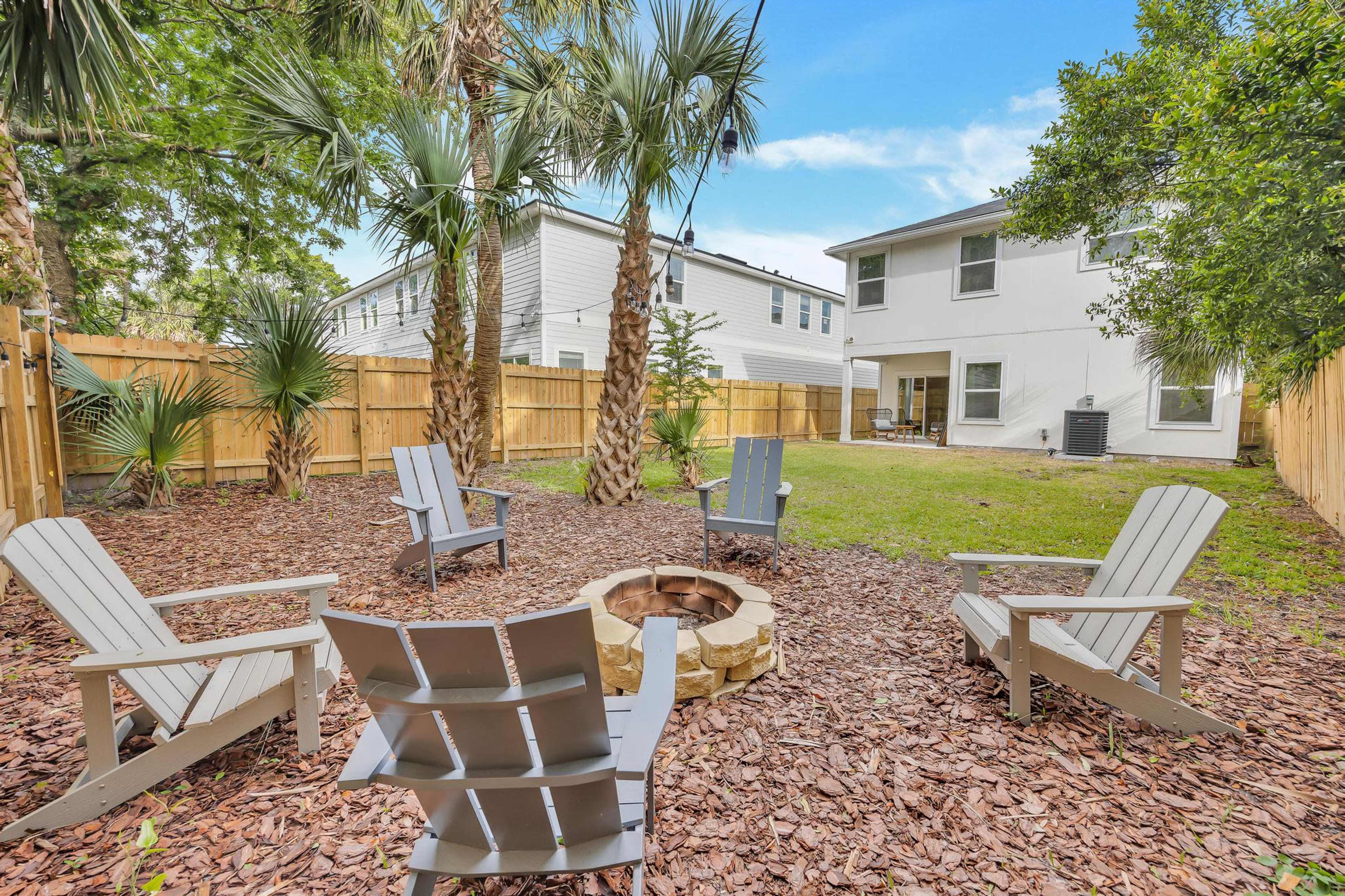 A circular fire pit surrounded by wooden Adirondack chairs is set in a backyard with grass and palm trees, enclosed by a wooden fence.