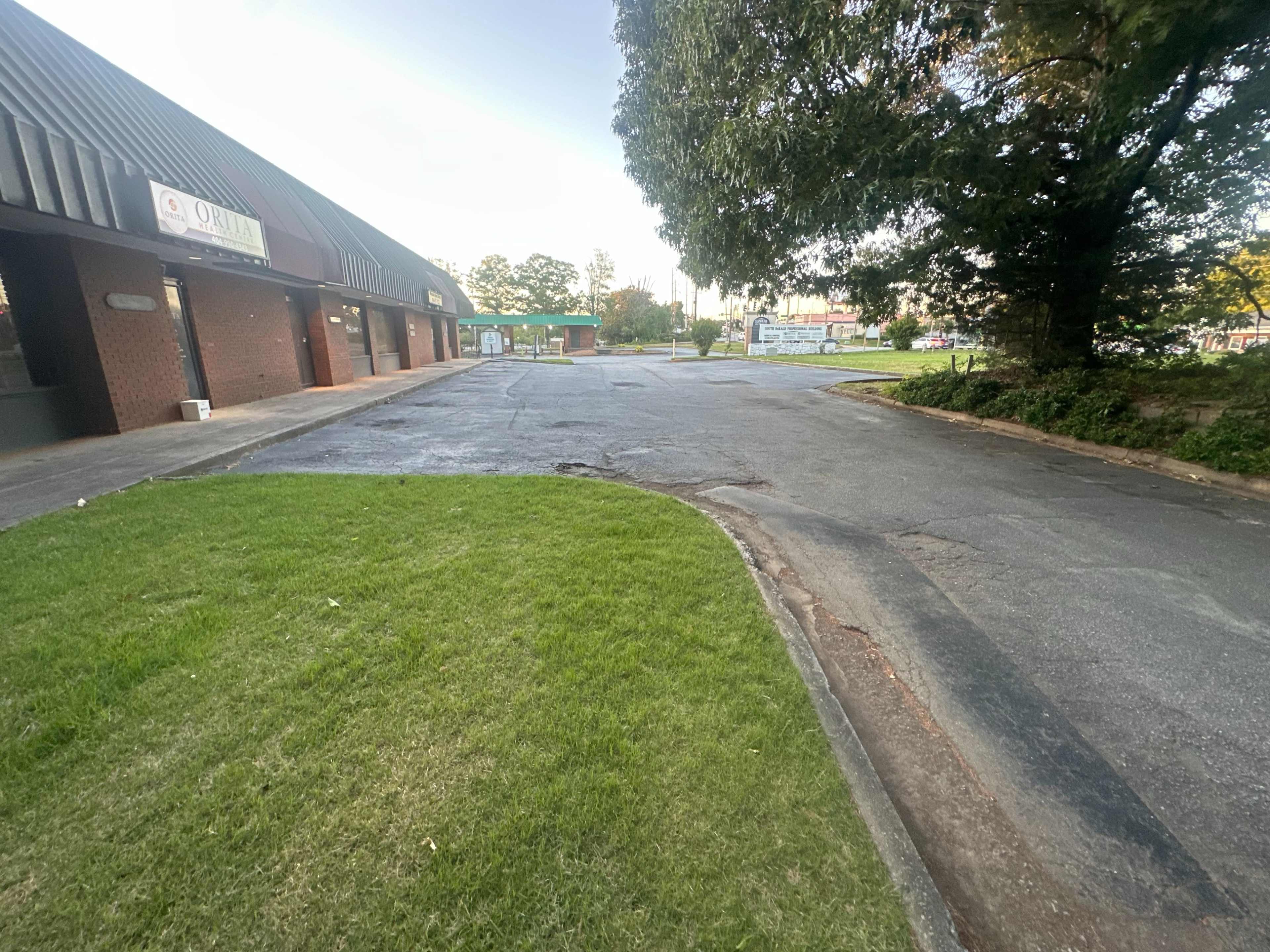 The image shows a paved area with a grassy edge, leading to a commercial building on the left and a sign in the distance on the right.