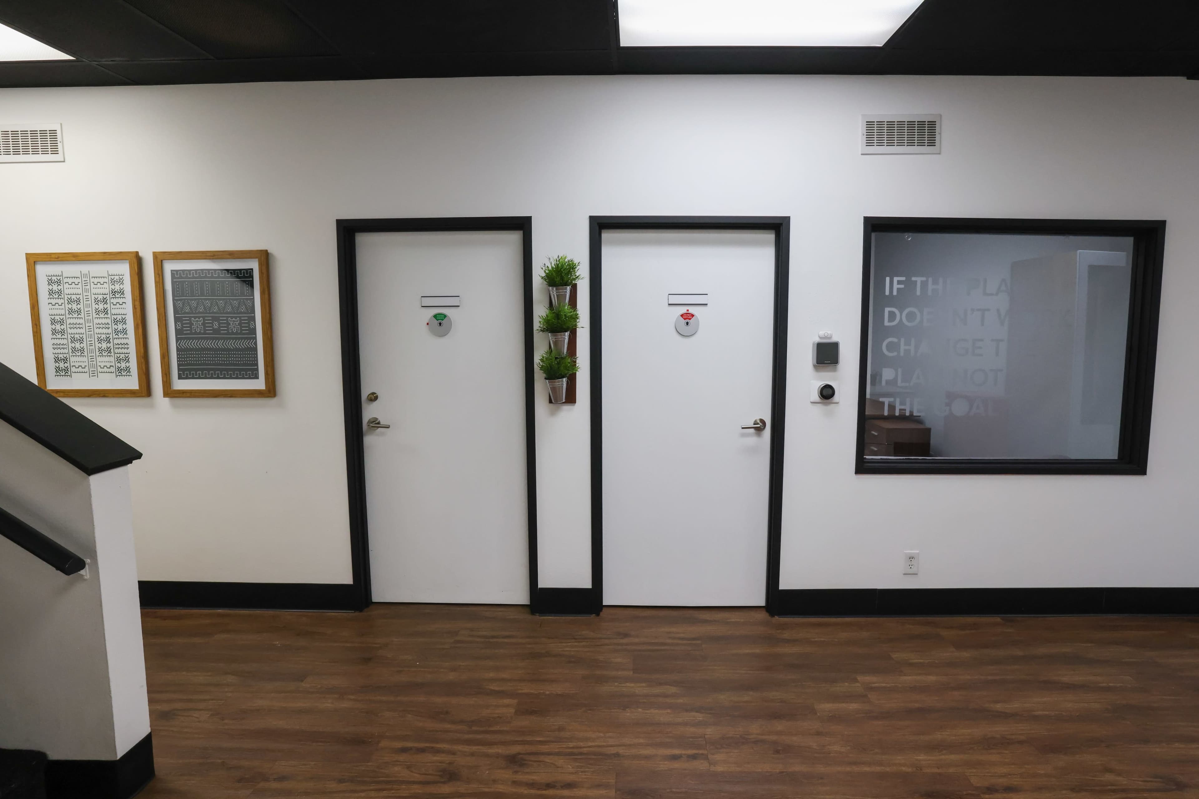 A corridor with two white doors labeled with red and green signs, alongside framed artwork on the wall and a set of planters.
