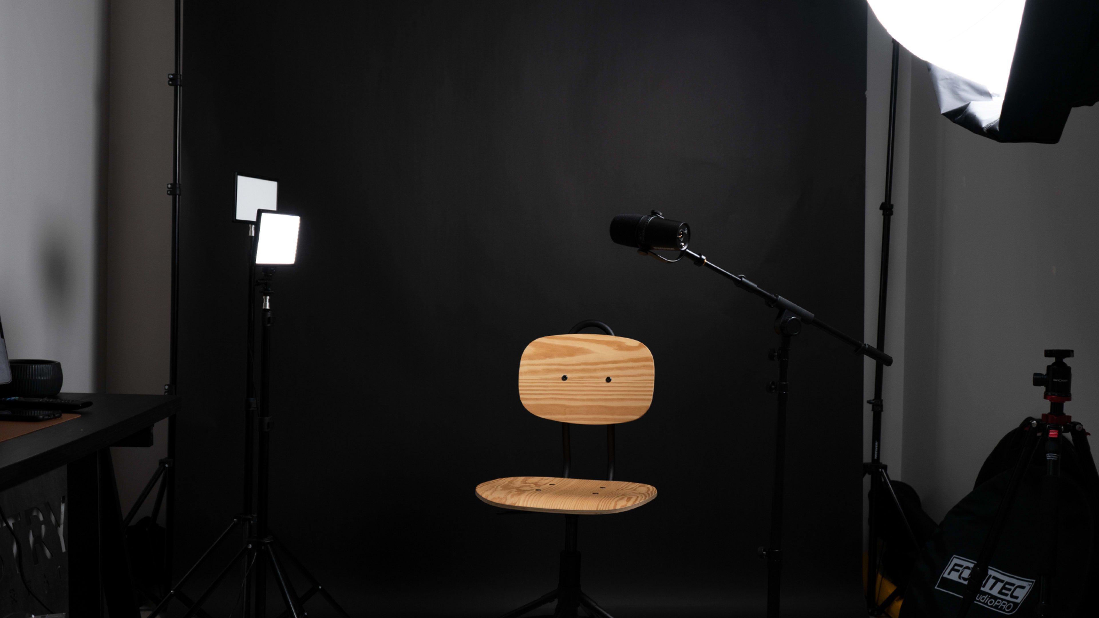 A wooden chair sits in front of a black backdrop, accompanied by a microphone and lighting equipment.