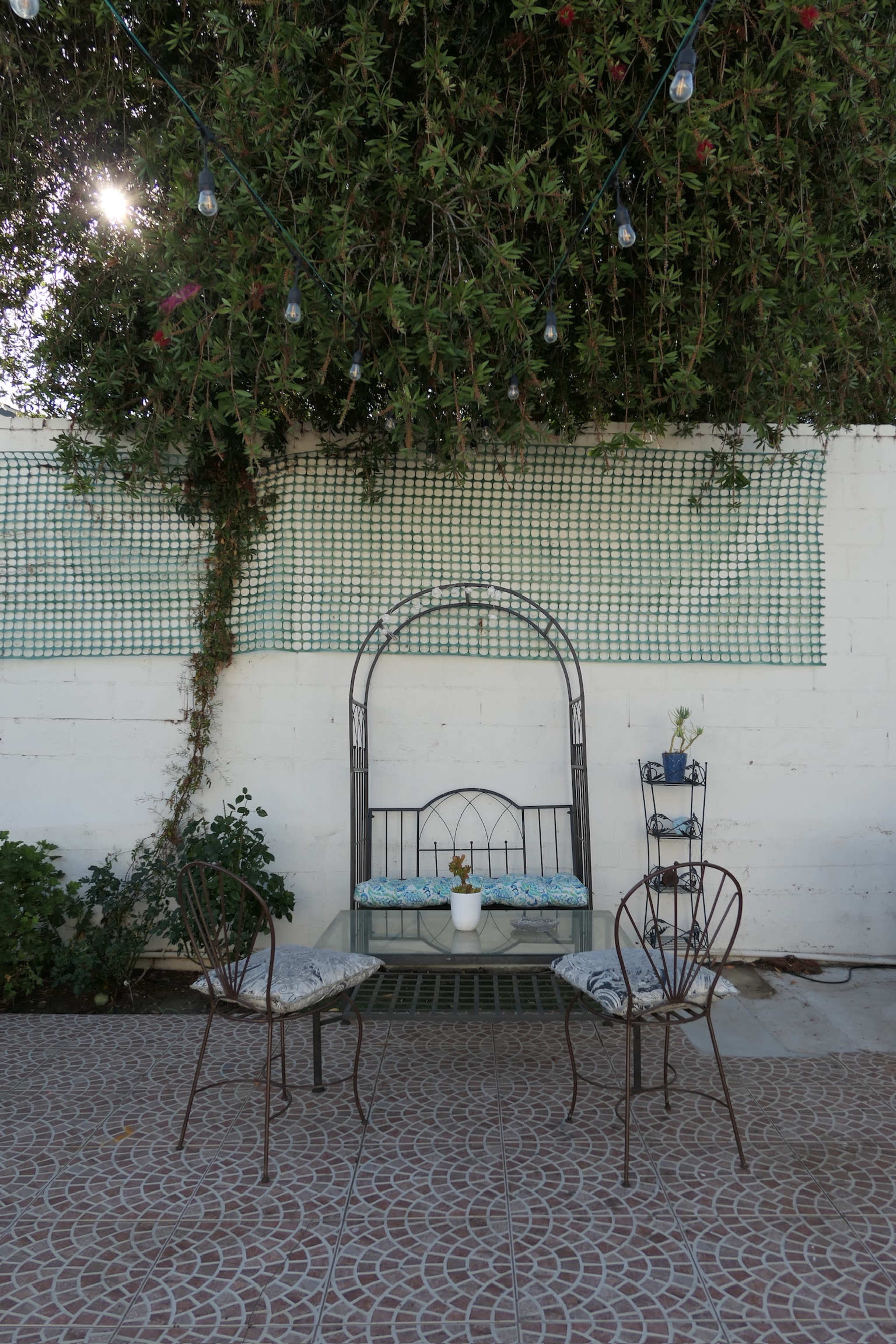 A metal bench with a potted plant sits against a white wall, flanked by two chairs on a tiled patio beneath string lights.