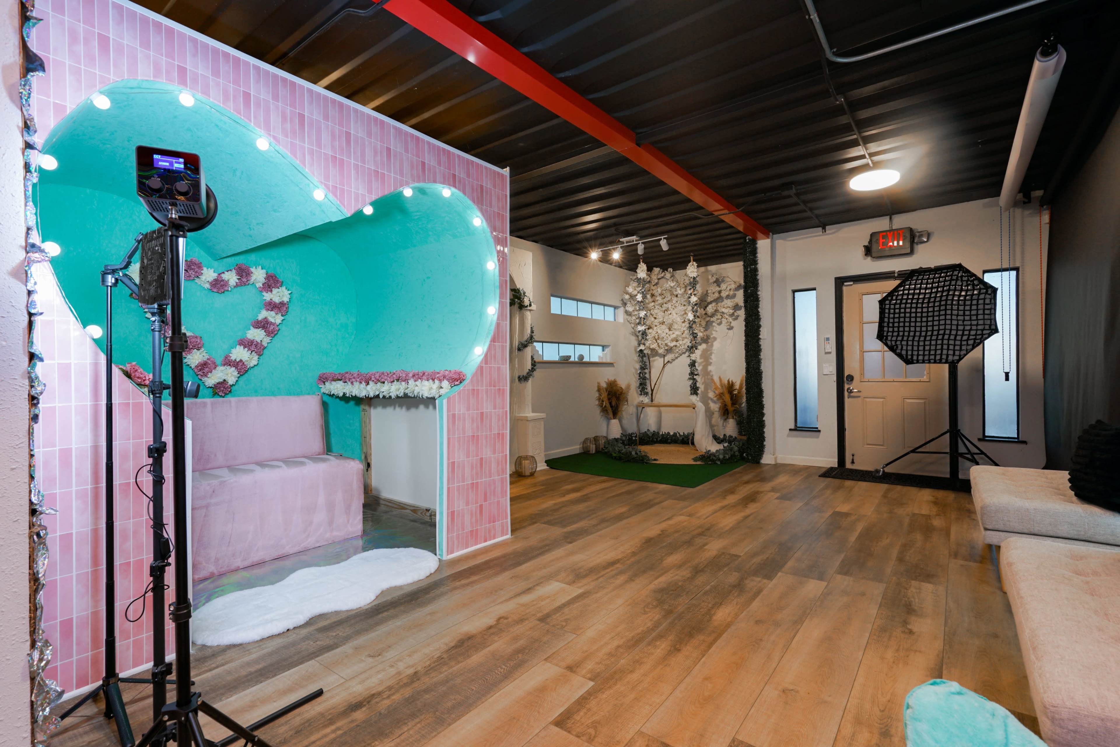 The image shows a modern photo studio with a heart-shaped backdrop, decorative lighting, and a combination of cozy seating and green accents.