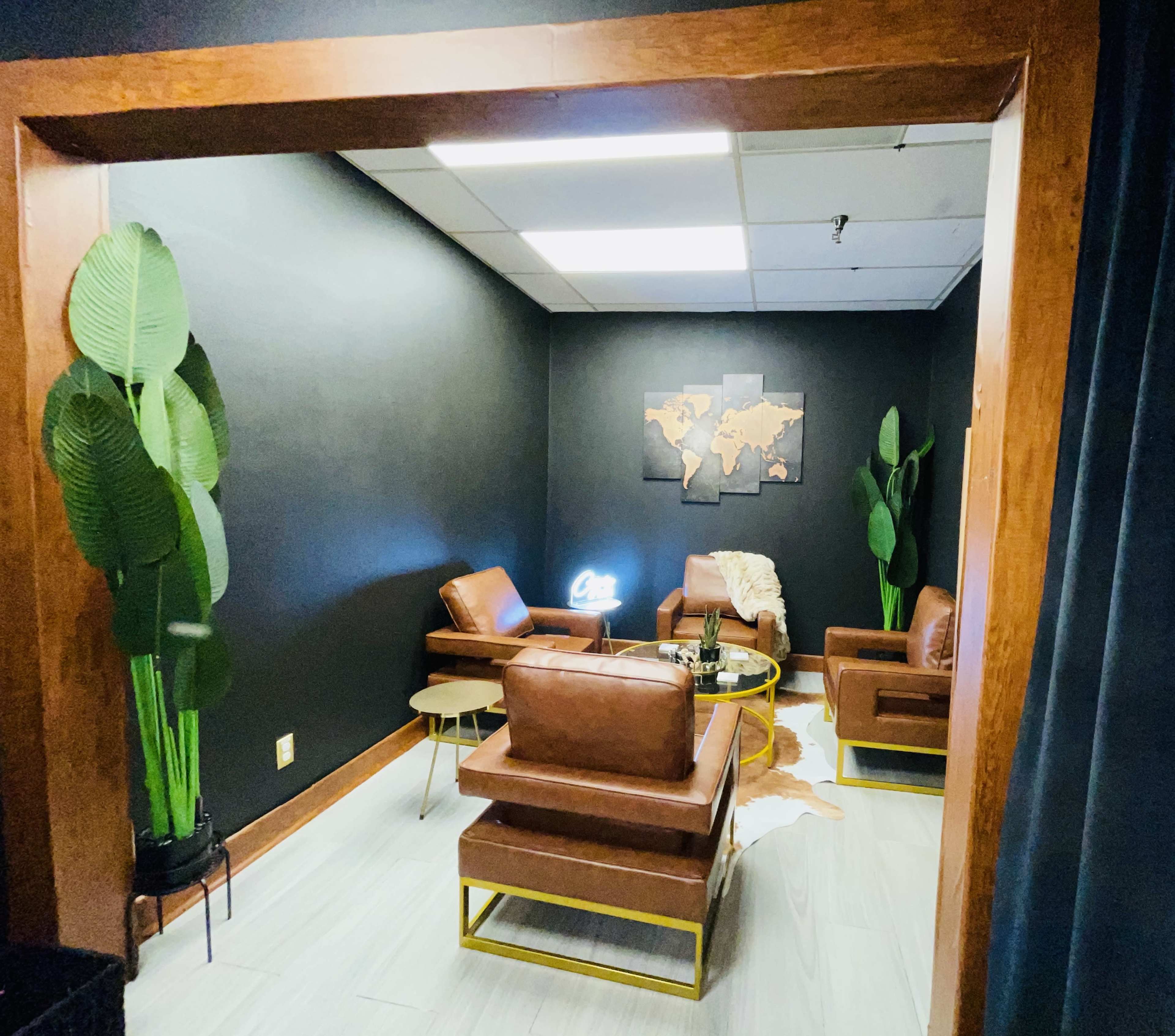 A modern lounge area features brown leather chairs, a small table, and a world map on the wall, framed by indoor plants and dark-colored walls.