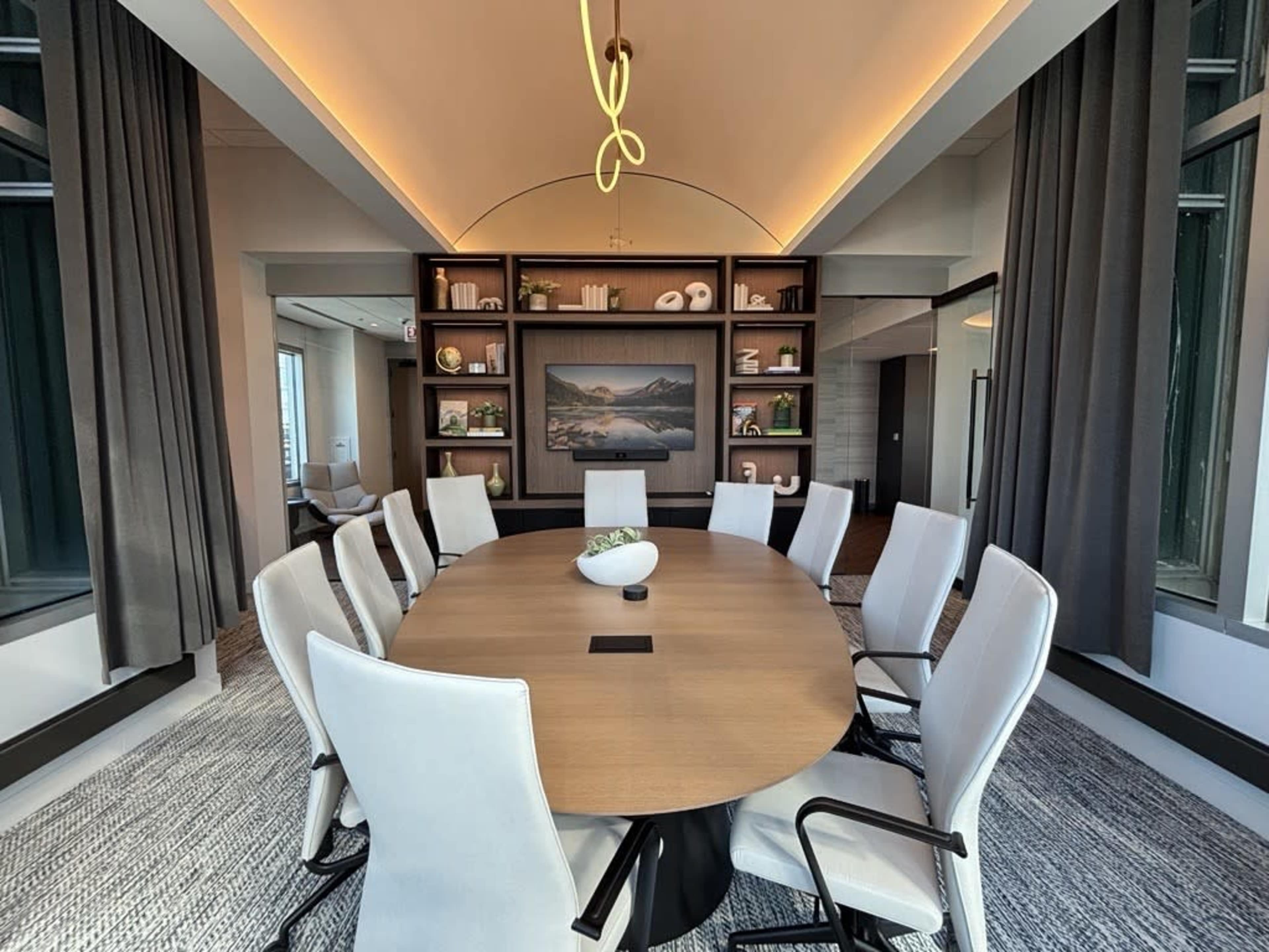 A modern conference room features a circular table surrounded by white chairs, with a backdrop of bookshelves and a landscape painting.