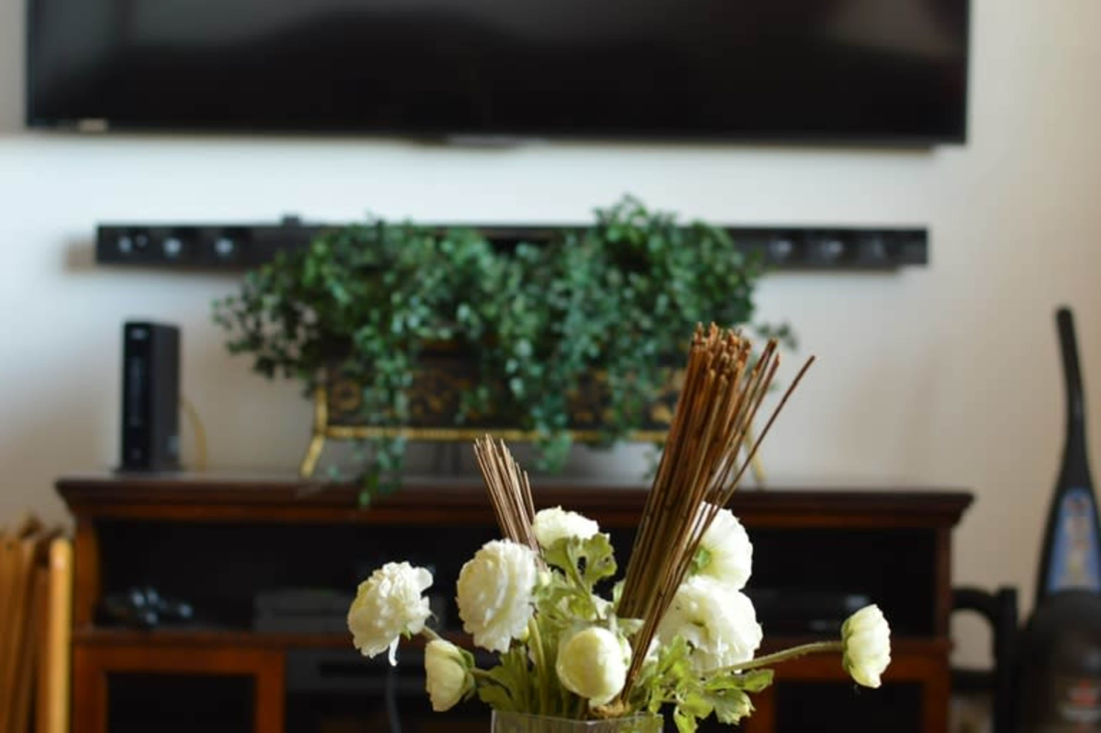 A vase with white flowers and decorative sticks sits on a table in front of a television and a stand with plants.