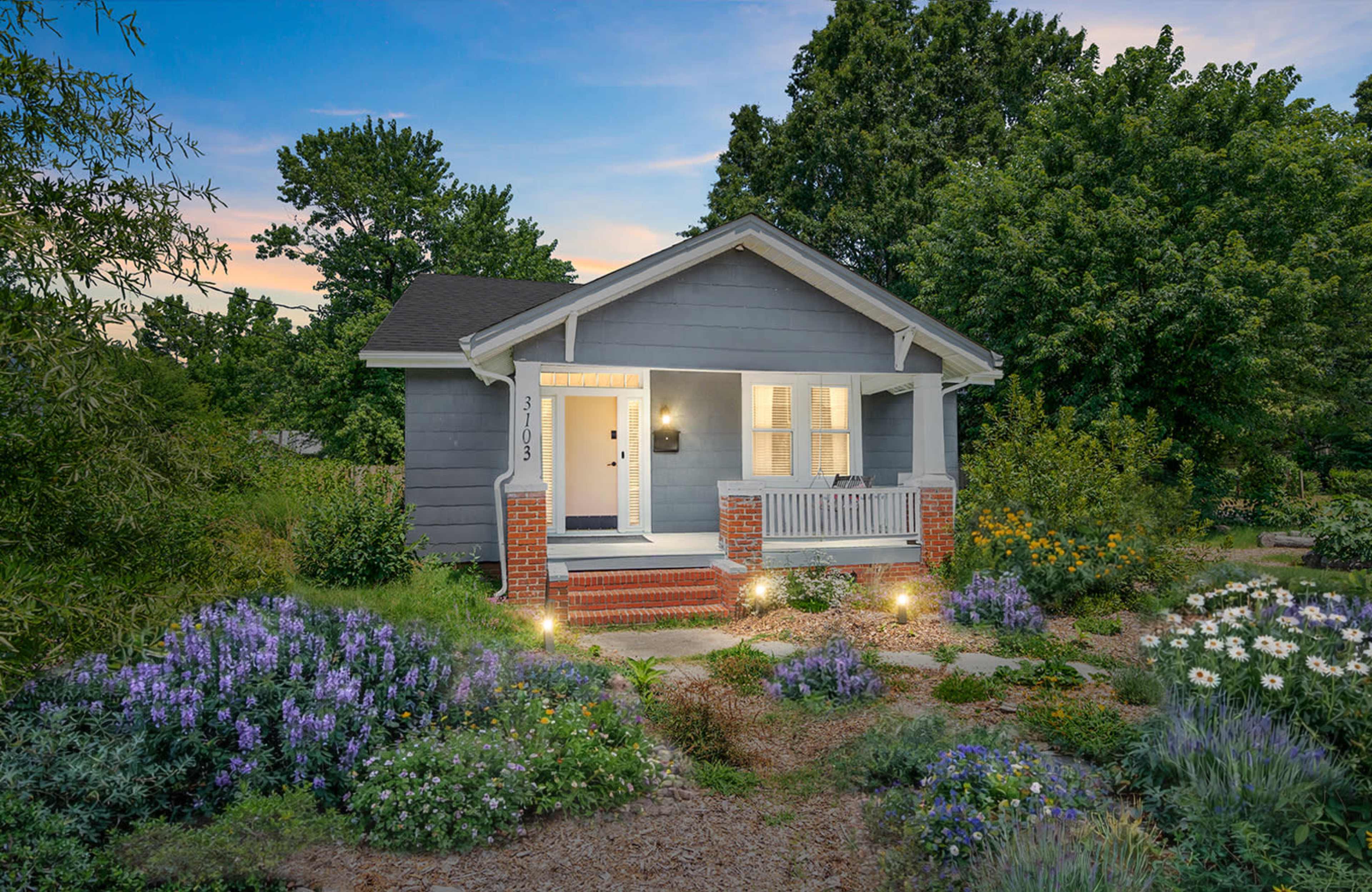 The image shows a small gray house with a front porch, surrounded by blooming flowers and lush greenery under a twilight sky.