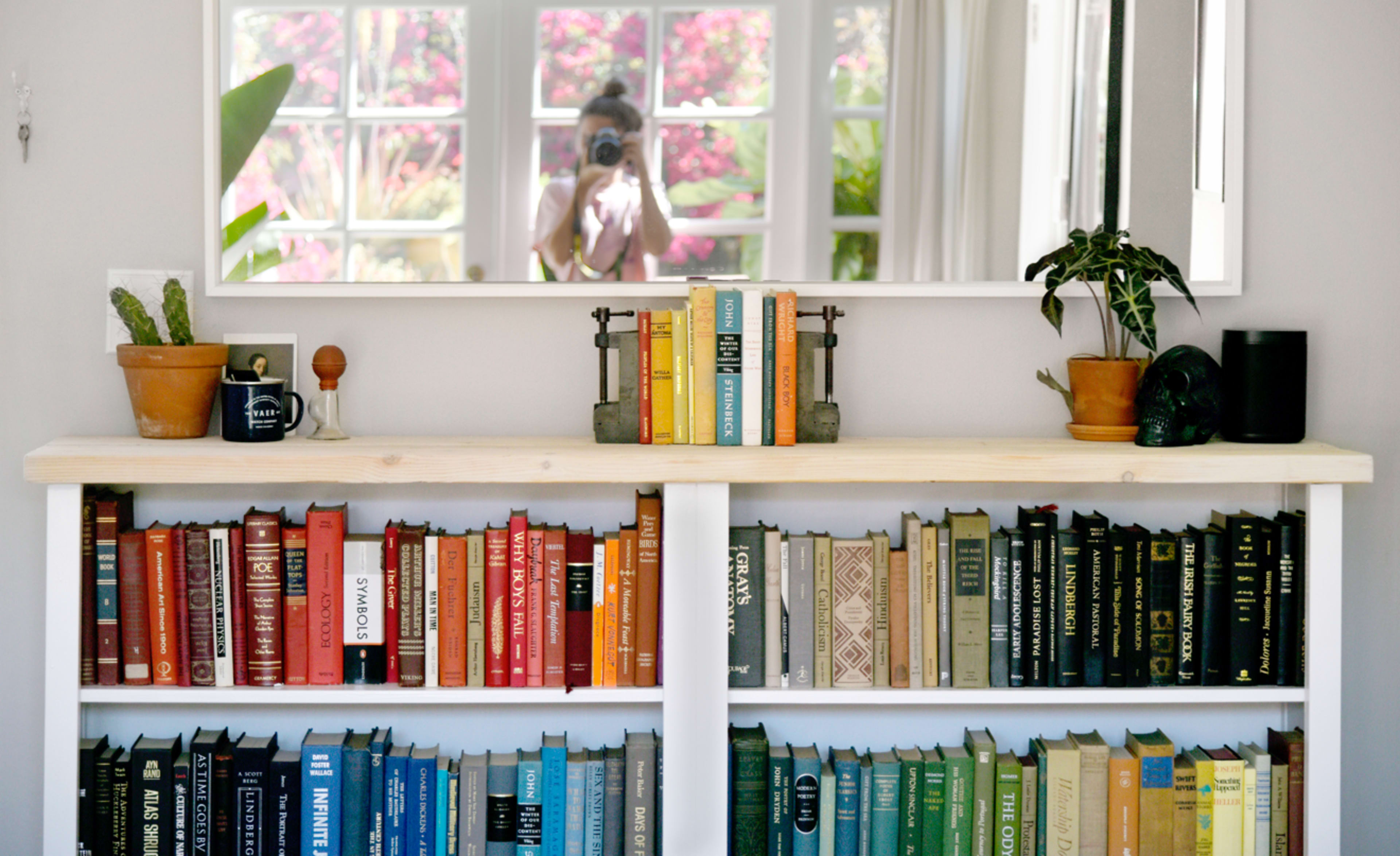 A bookshelf with colorful books and decorative plants is placed under a mirror reflecting a person taking a photo.