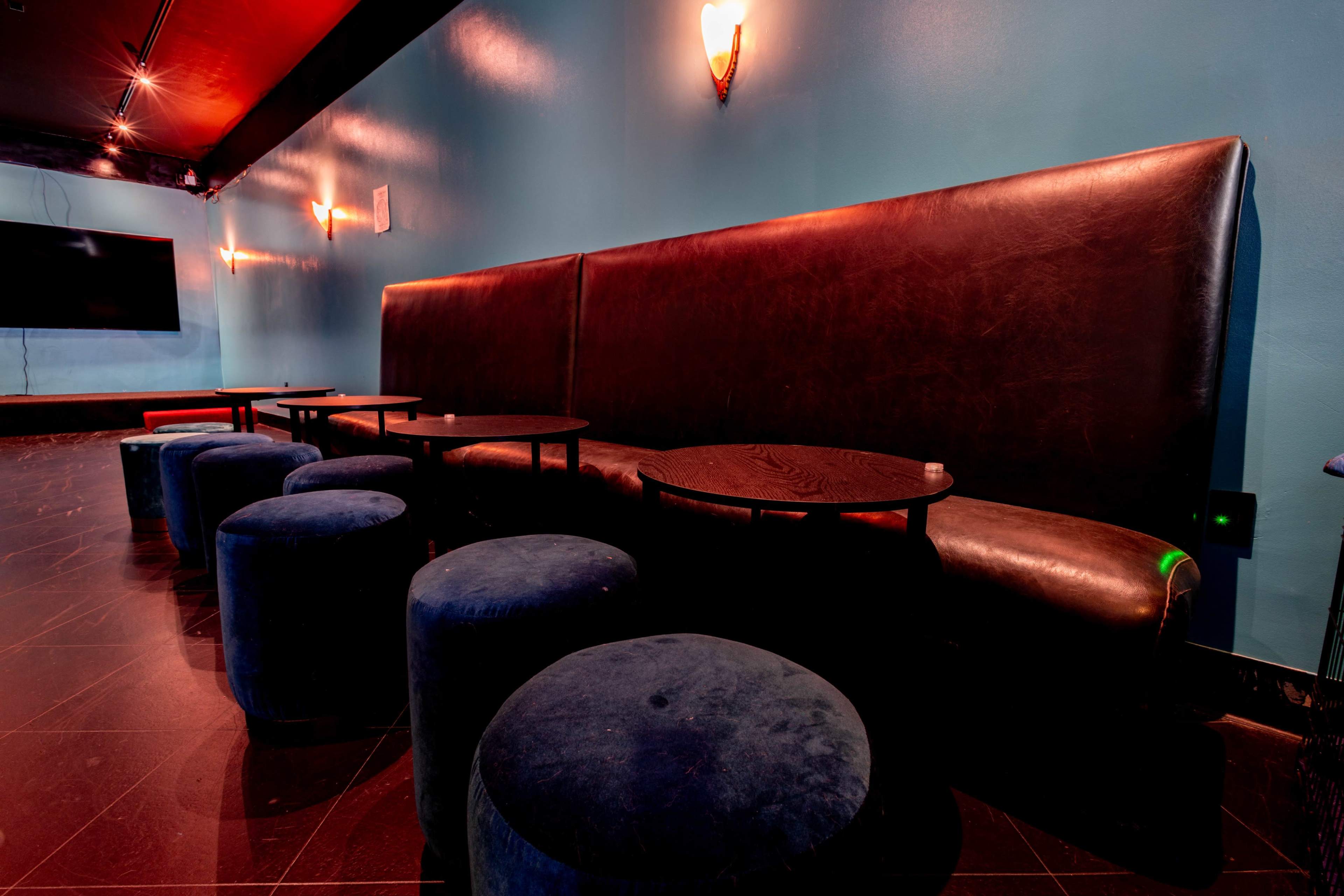 A long leather bench is paired with circular blue stools and small dark tables against a teal wall in a dimly lit interior space.