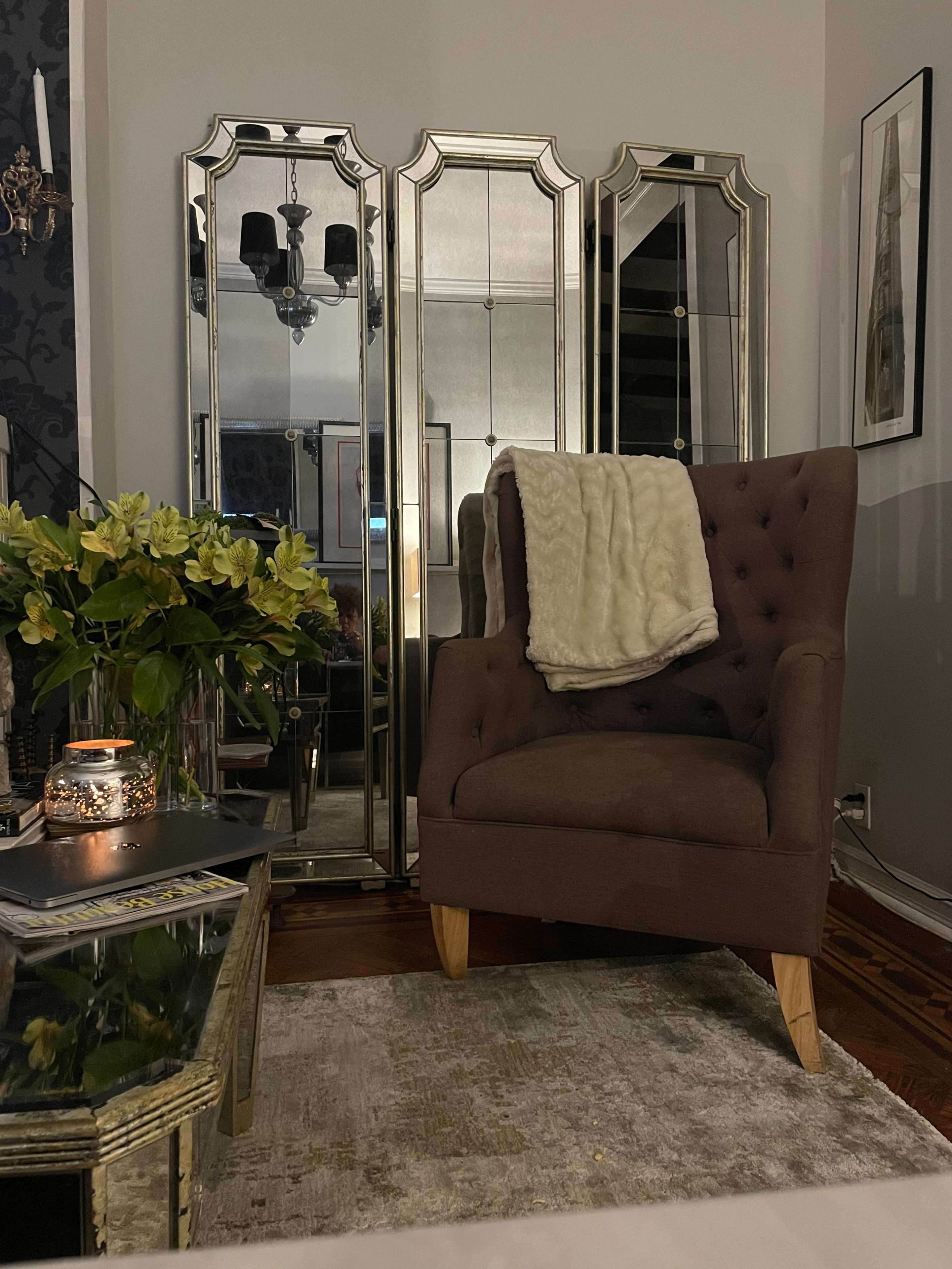 A brown tufted armchair is positioned near a mirrored folding screen, accompanied by a coffee table adorned with flowers and a lit candle.