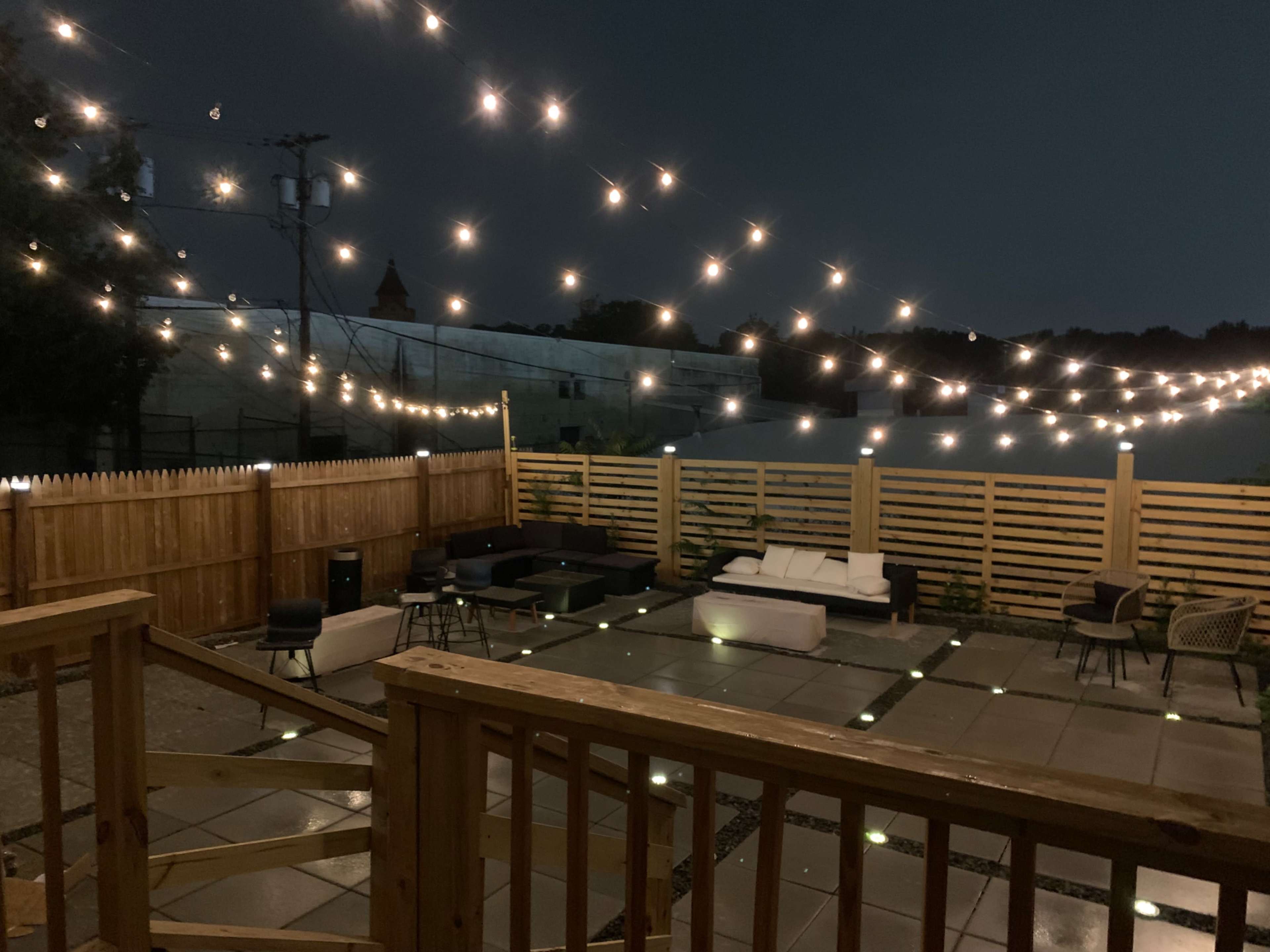 A patio area features a stone floor illuminated by string lights overhead, surrounded by wooden fencing and various seating arrangements.
