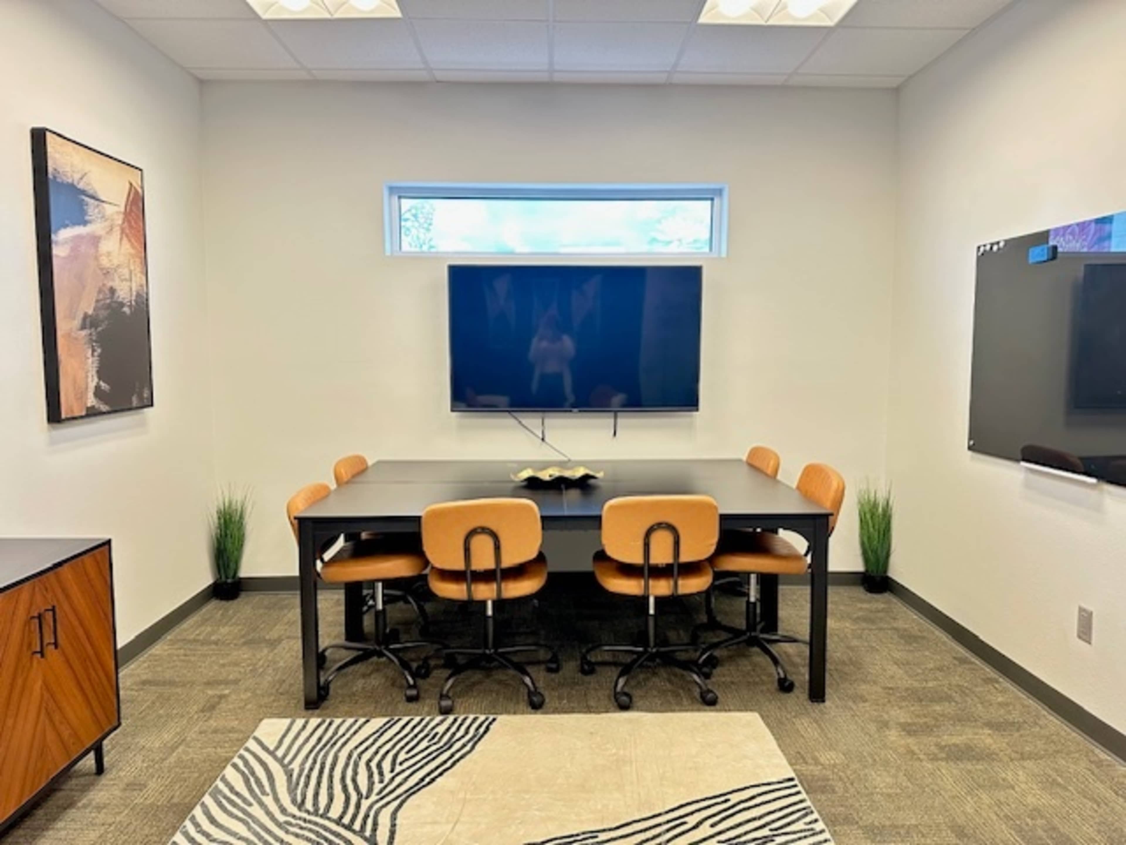 A modern conference room features a large table surrounded by orange chairs, with a television mounted on the wall and artwork on display.