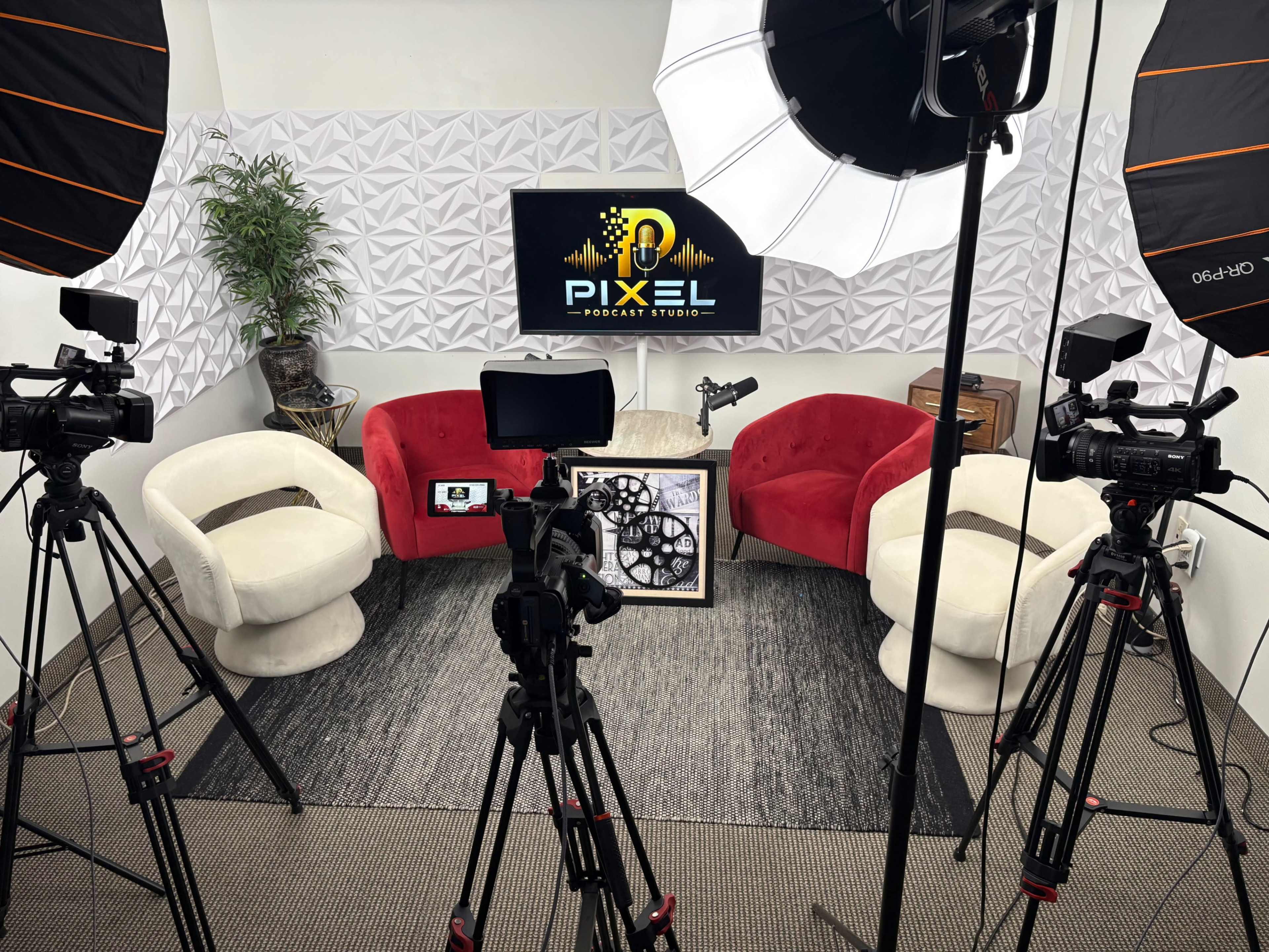 The image shows a podcast studio with two red and white chairs, several cameras on tripods, a light setup, and a television displaying the Pixel Podcast Studio logo in front of a textured wall.