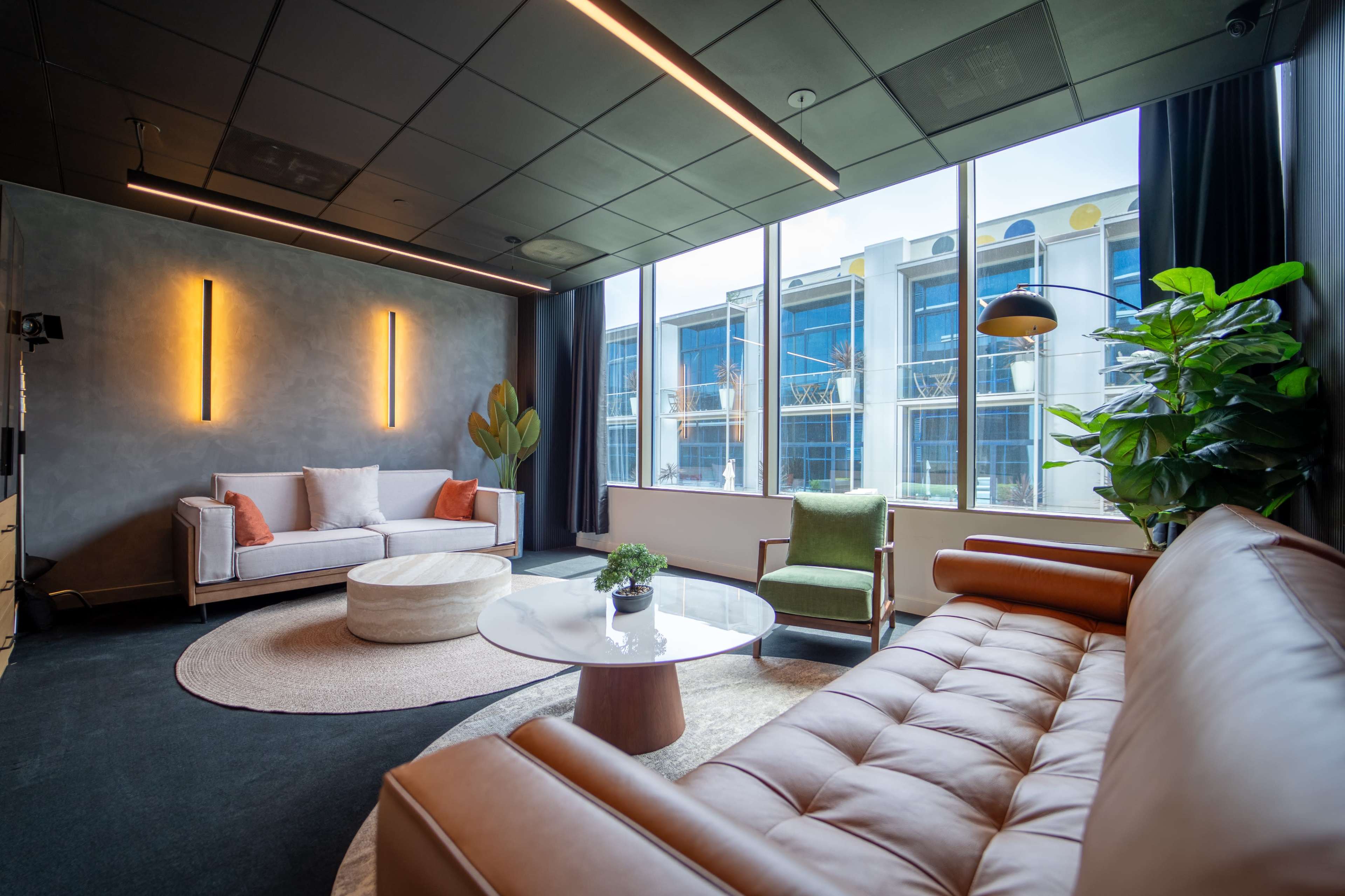 The image shows a modern lounge with a beige leather sofa, a round coffee table, a green accent chair, and large windows revealing an outdoor view.
