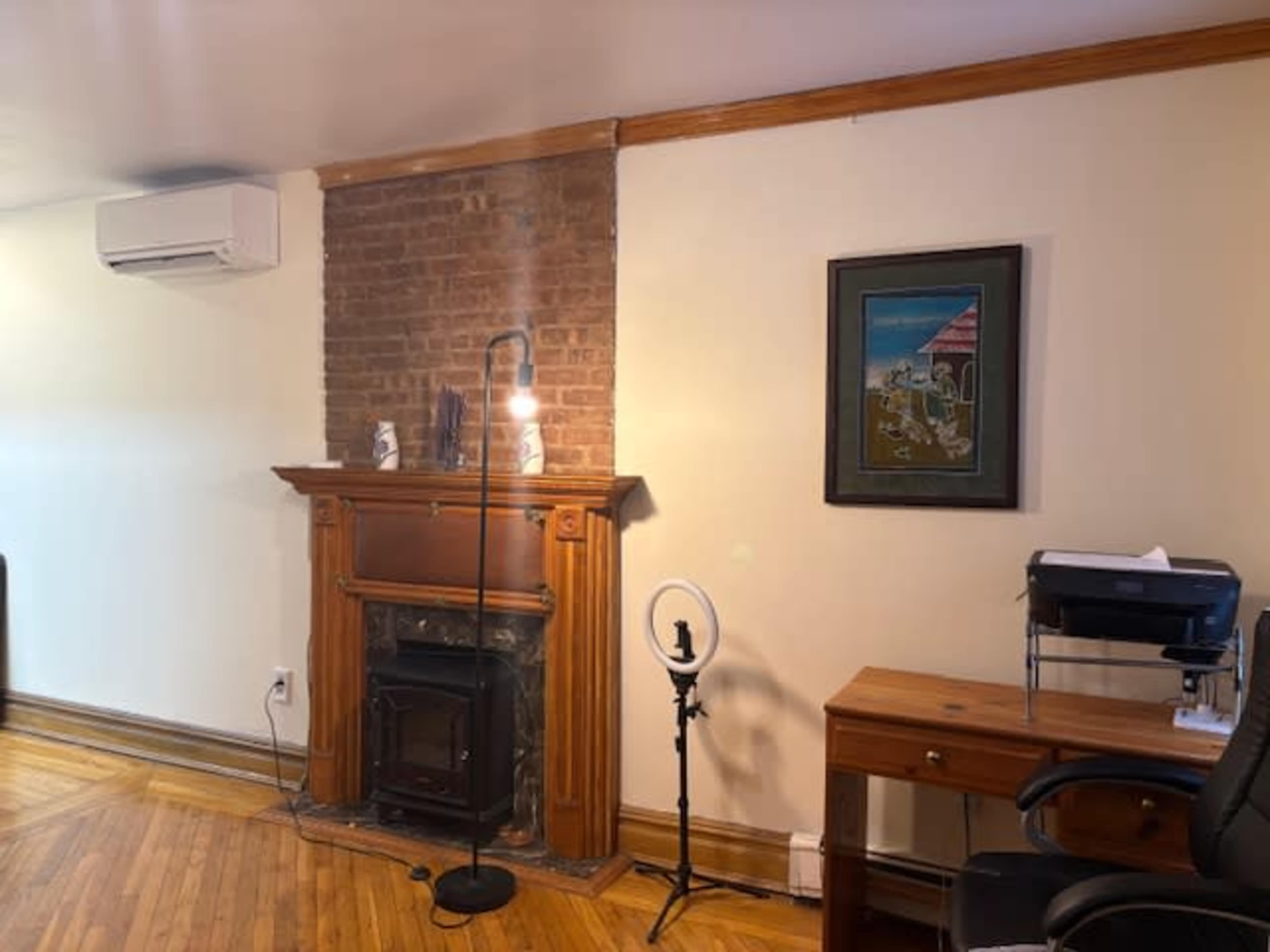 The image shows a room with a decorative brick wall, a wooden fireplace mantel, an art piece on the wall, a floor lamp, and a desk with an office chair.