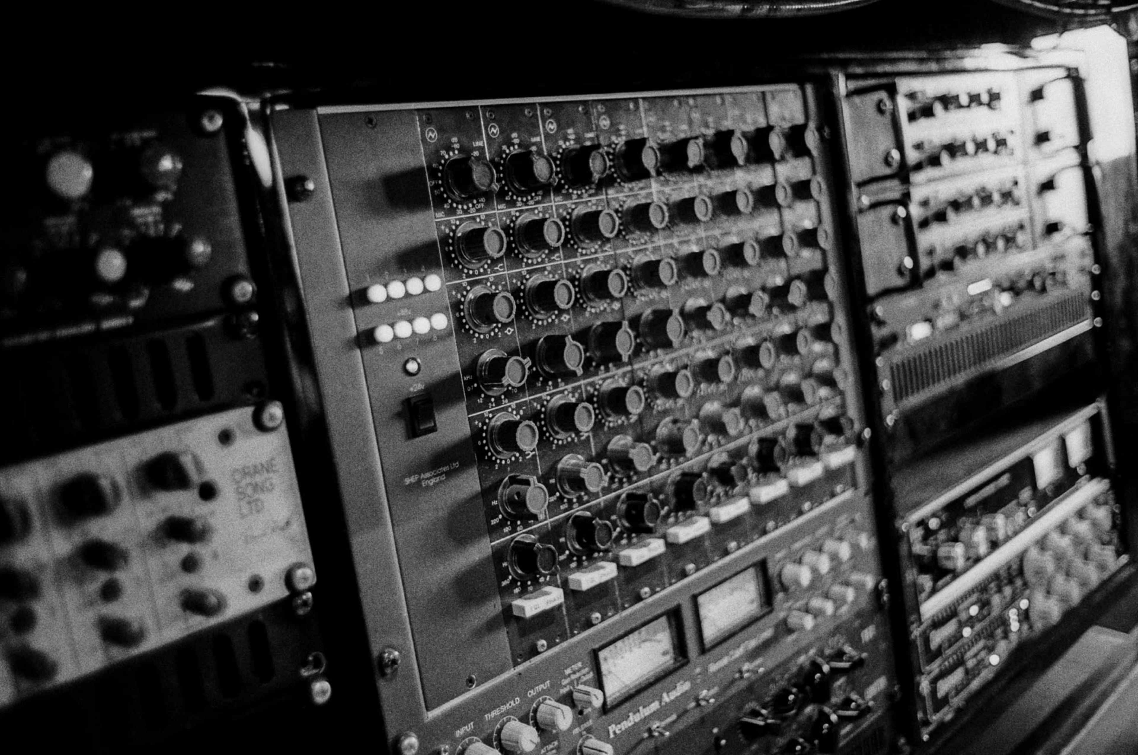 The image shows a close-up view of various audio equipment and mixing consoles featuring numerous knobs and sliders.