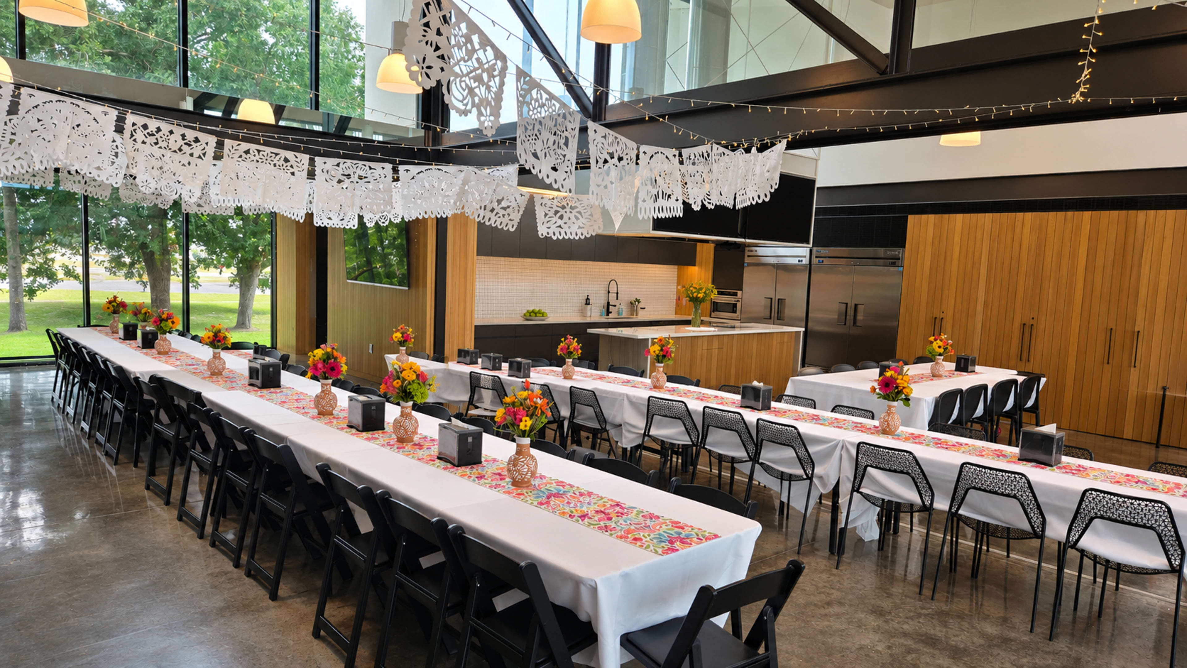 The image shows a bright, modern event space set up with long tables adorned with colorful table runners and floral centerpieces, surrounded by large windows overlooking greenery.