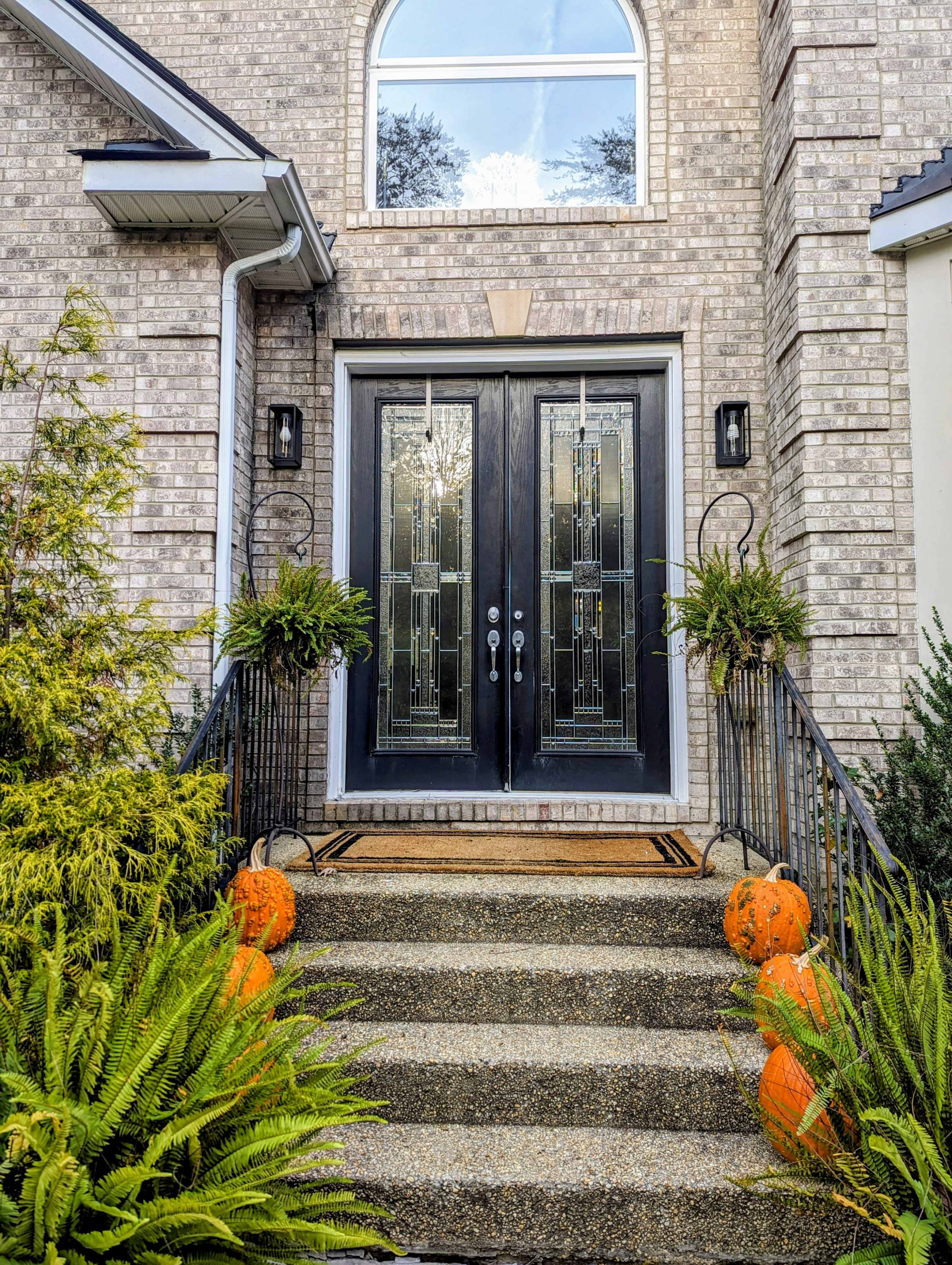 The entrance features a pair of black double doors with decorative glass, flanked by potted ferns and orange pumpkins on the steps.