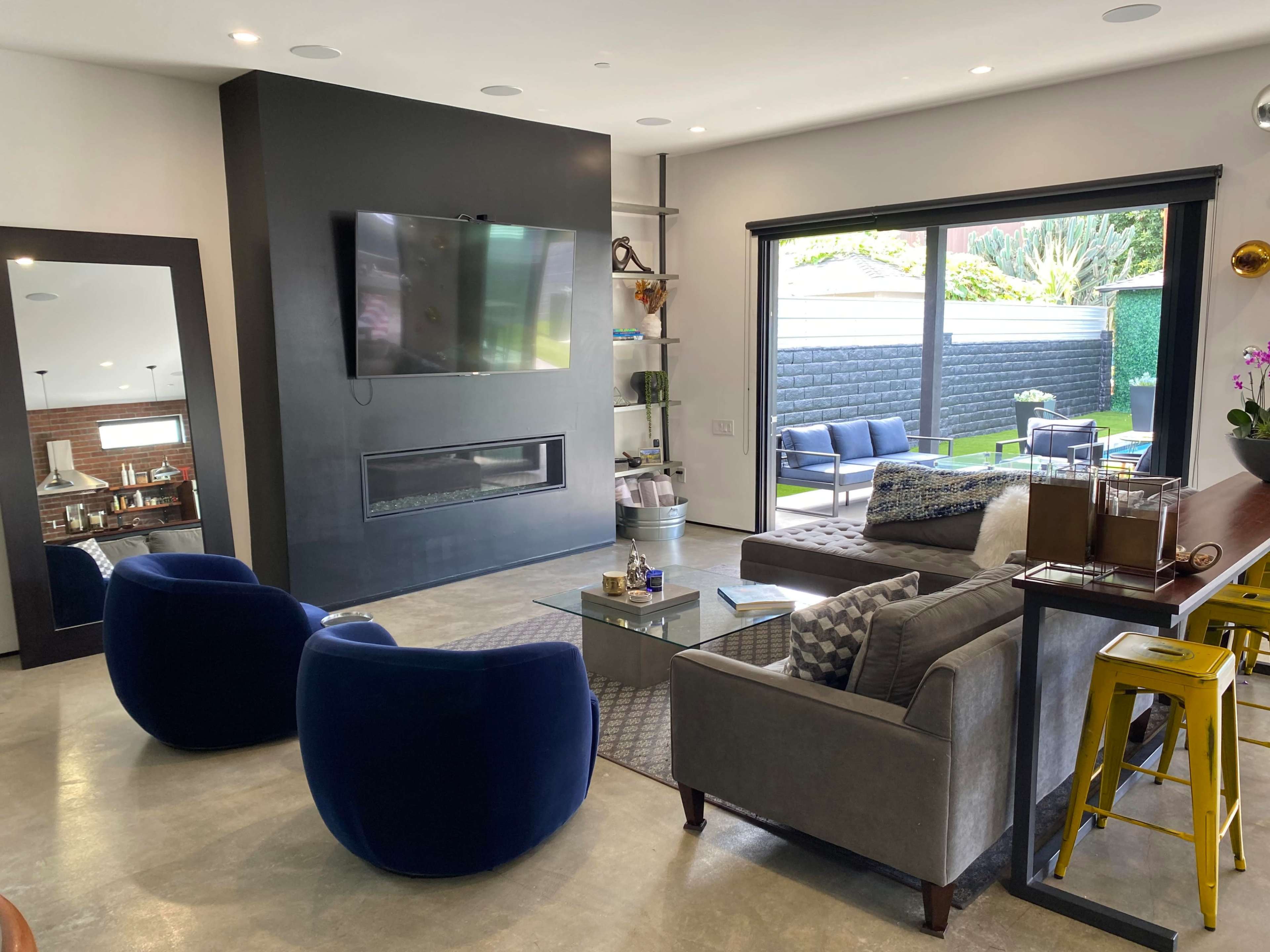 The modern living room features a television mounted on a black wall, a gray sofa, blue accent chairs, and large sliding glass doors leading to a patio area.