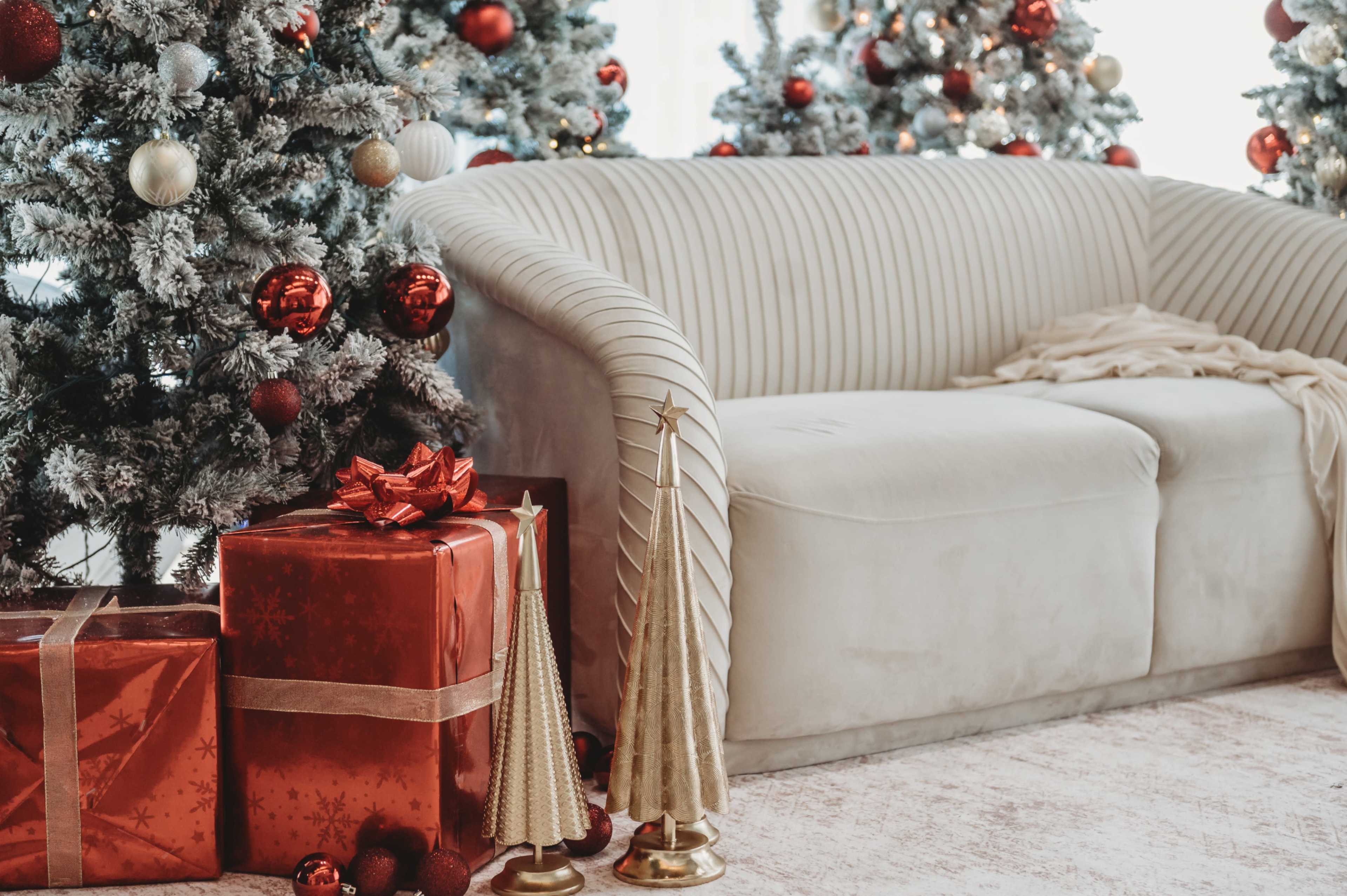 The scene features a cozy, light-colored sofa surrounded by decorated Christmas trees and wrapped gifts in red and gold.