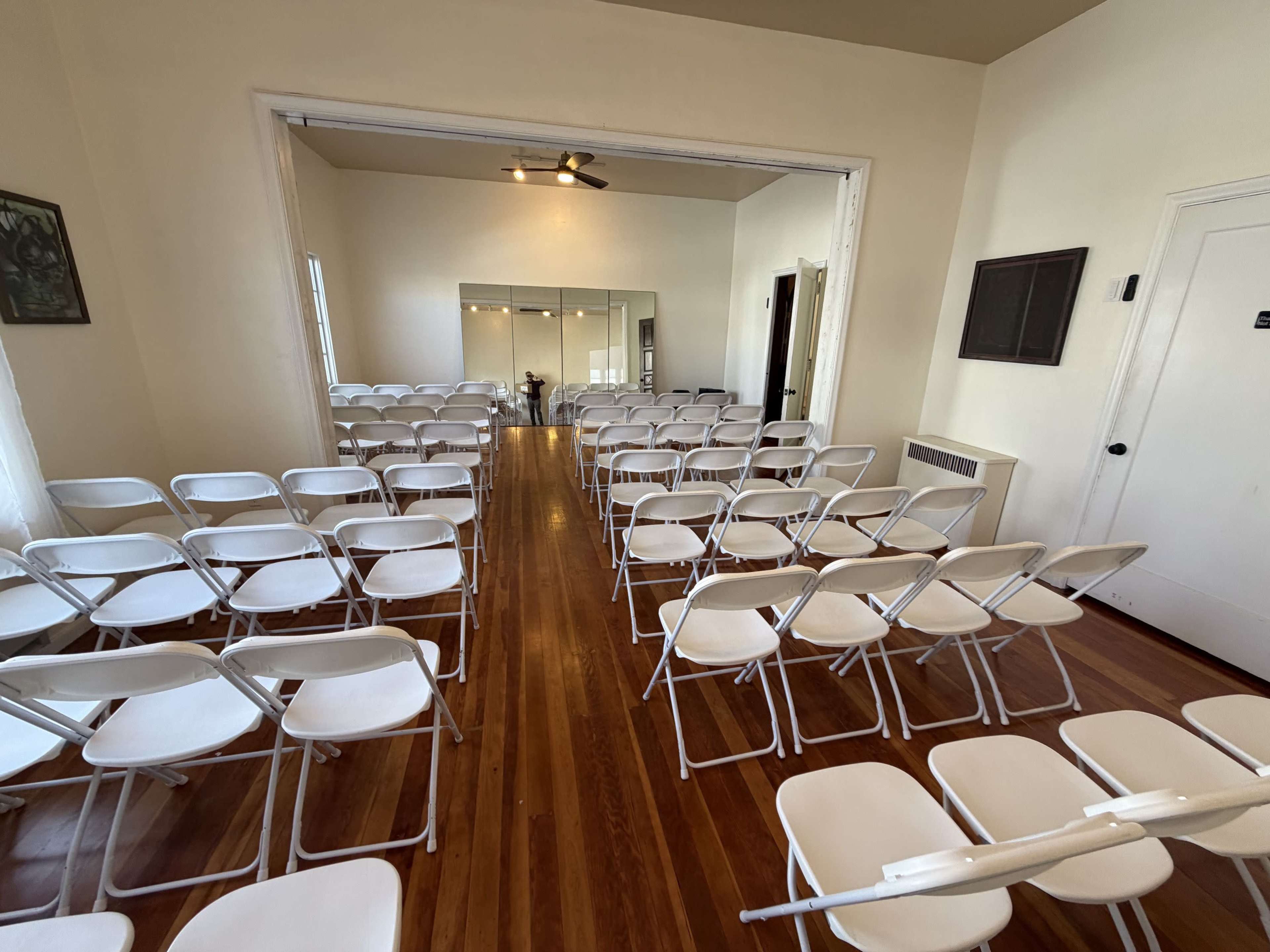 The image shows a room set up with rows of white folding chairs facing a mirror and a door, with wooden flooring and neutral-colored walls.