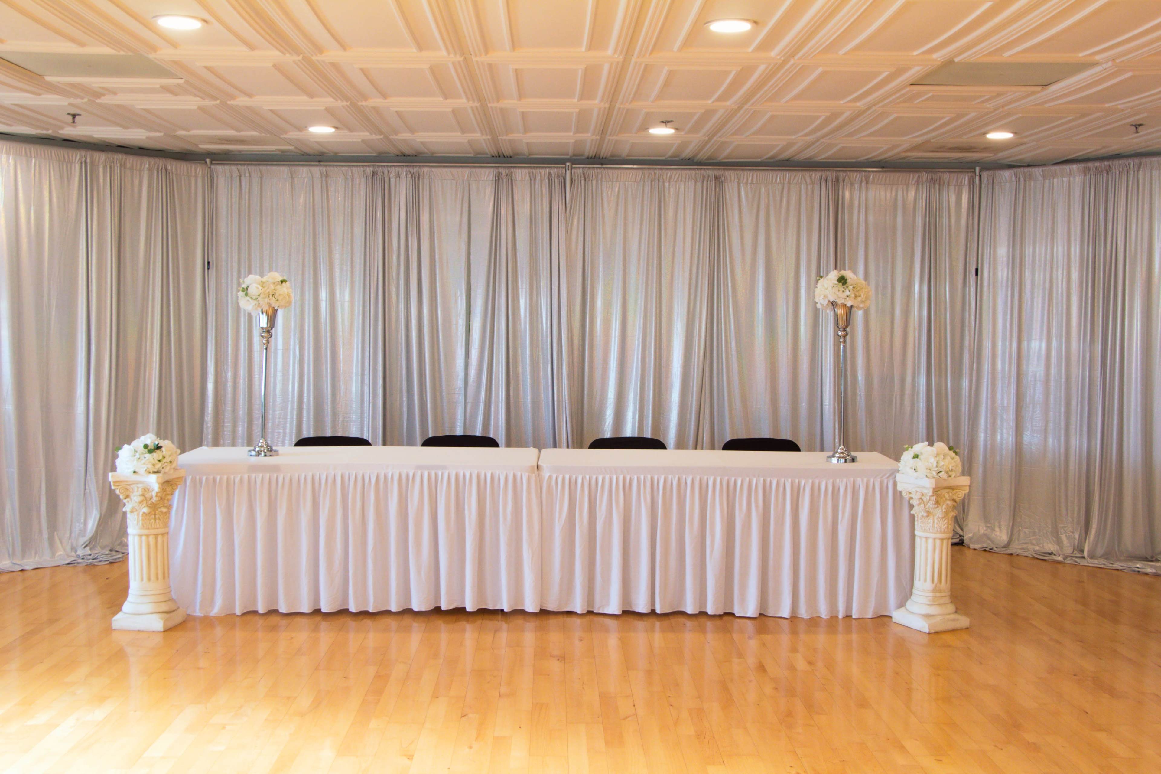 The image shows a decorated event space with a long table draped in white fabric, flanked by tall floral arrangements on pedestals and covered by silver drapery in the background.