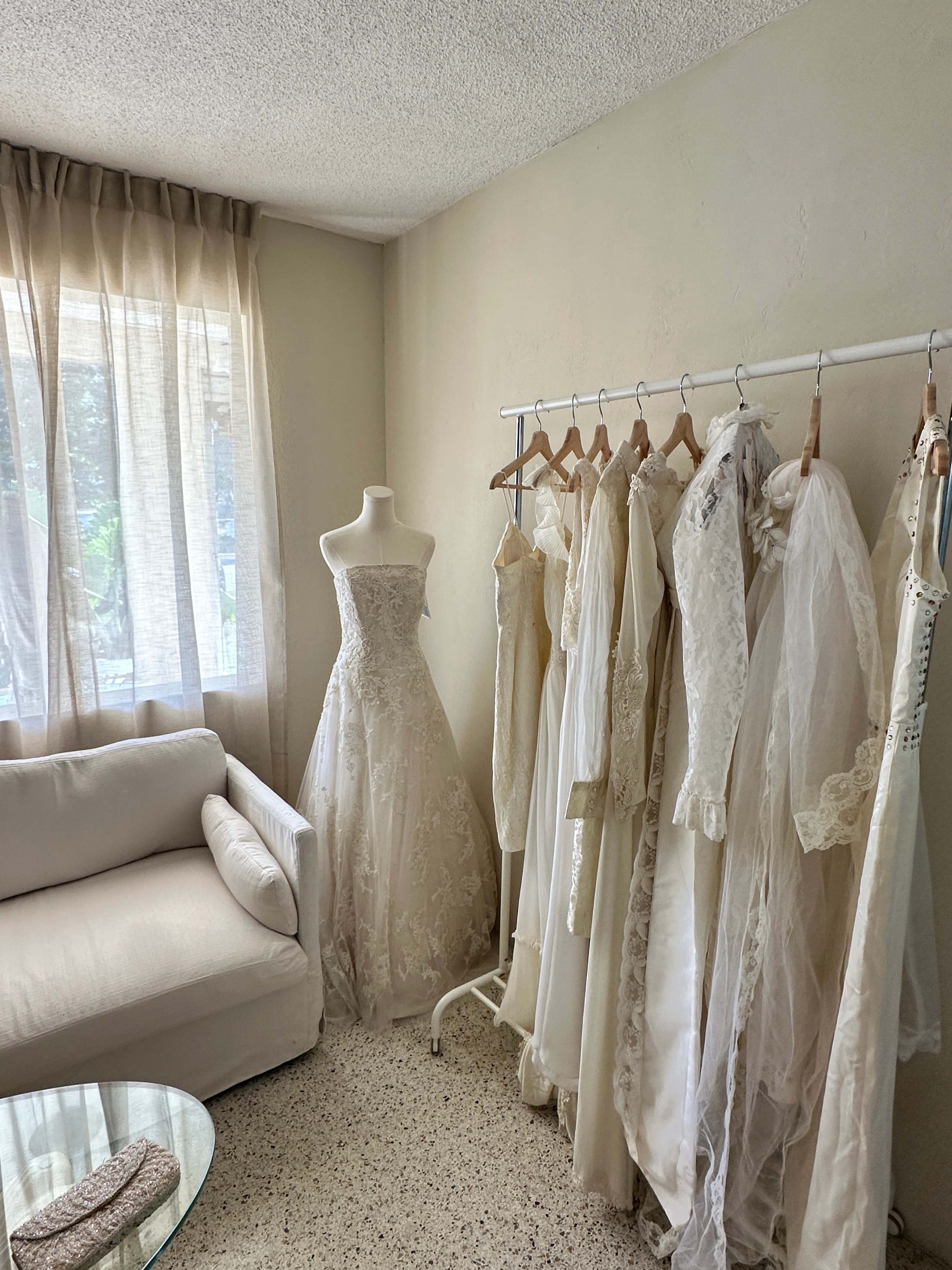 A sunlight-filled room features a mannequin dressed in a white gown alongside a rack displaying various white dresses and a beige sofa.