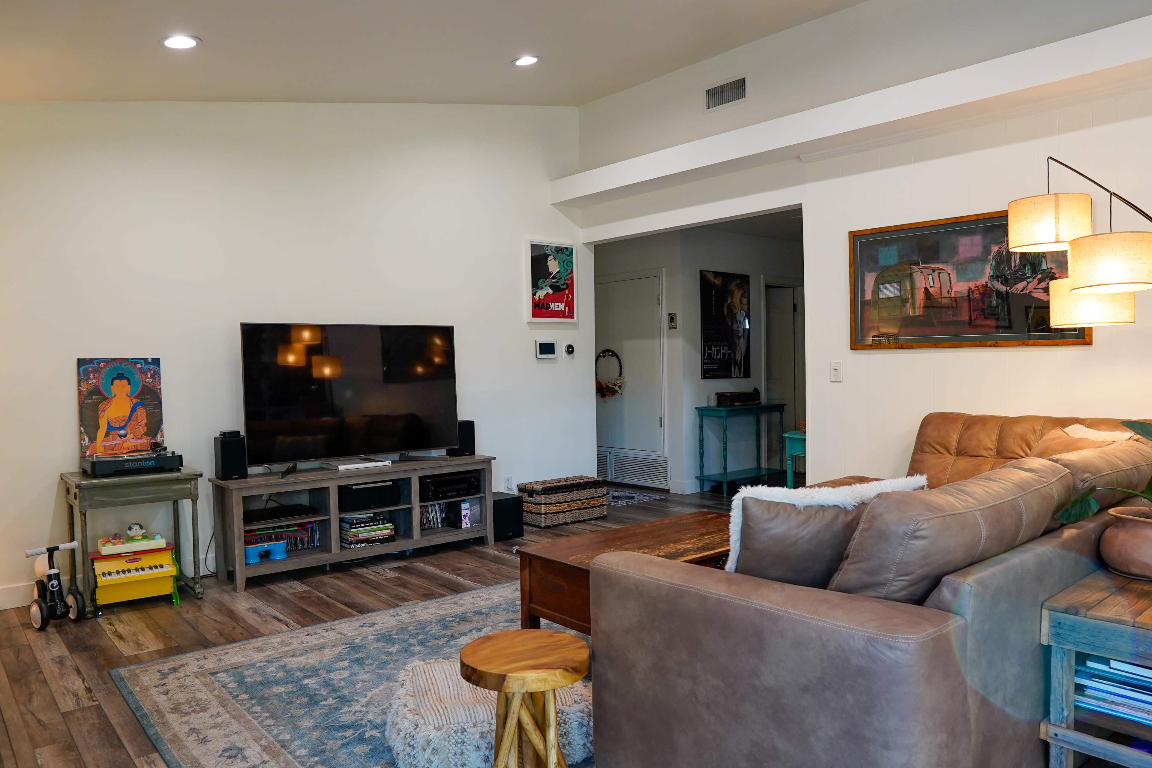 The image shows a cozy living room with a large television, a leather sectional sofa, a wooden coffee table, and various artworks on the walls.