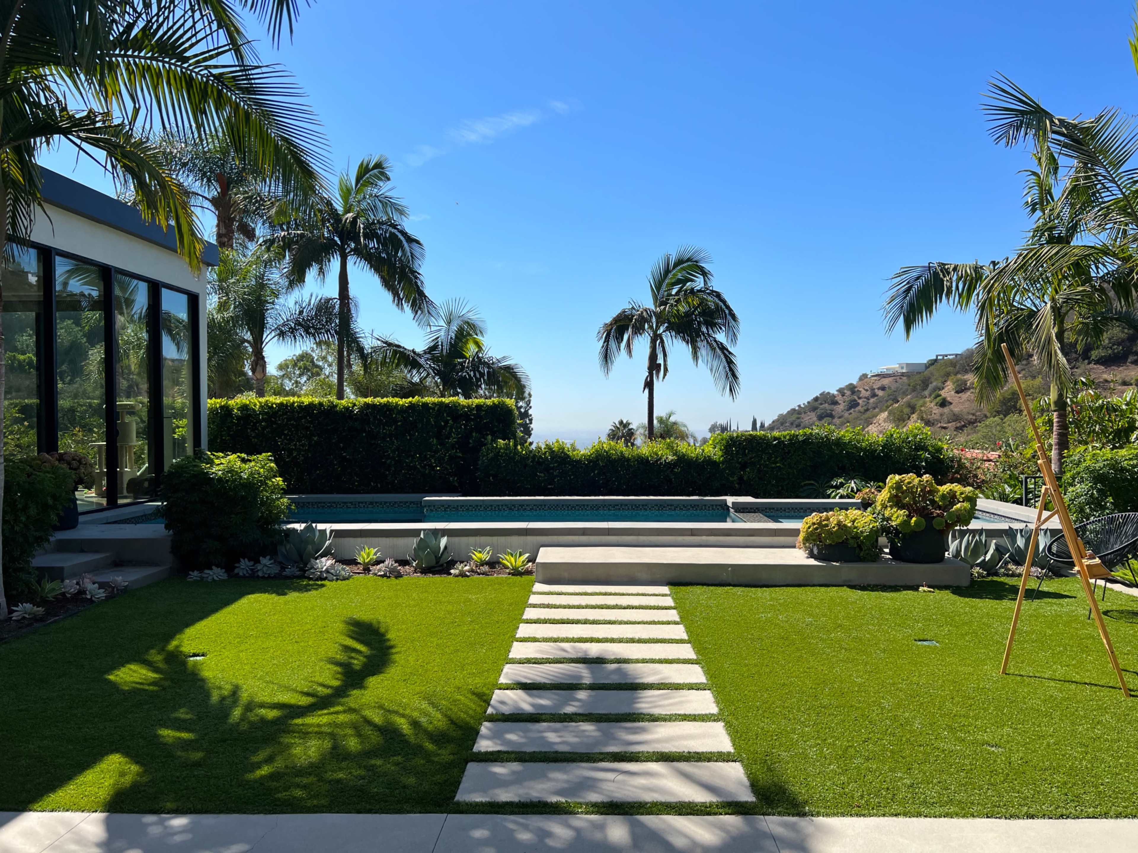 A modern backyard with a swimming pool, landscaped greenery, palm trees, and a clear blue sky.